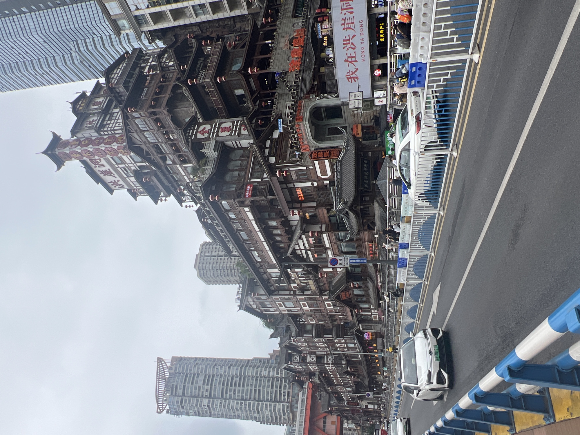 The image shows a street scene in Yuzhong District, Chongqing, China, featuring the Hongyadong complex, a distinctive multi-story building with traditional Chinese architecture. The building is adorned with wooden balconies, red lanterns, and tiered roofs, standing in contrast to the modern skyscrapers visible in the background. A white sedan is driving on the road in the foreground, which is separated from the sidewalk by a blue and white barrier. Several pedestrians are visible on the elevated walkways and in front of the buildings. The sky is overcast, indicating a cloudy day. Signs in Chinese characters are displayed on the buildings.