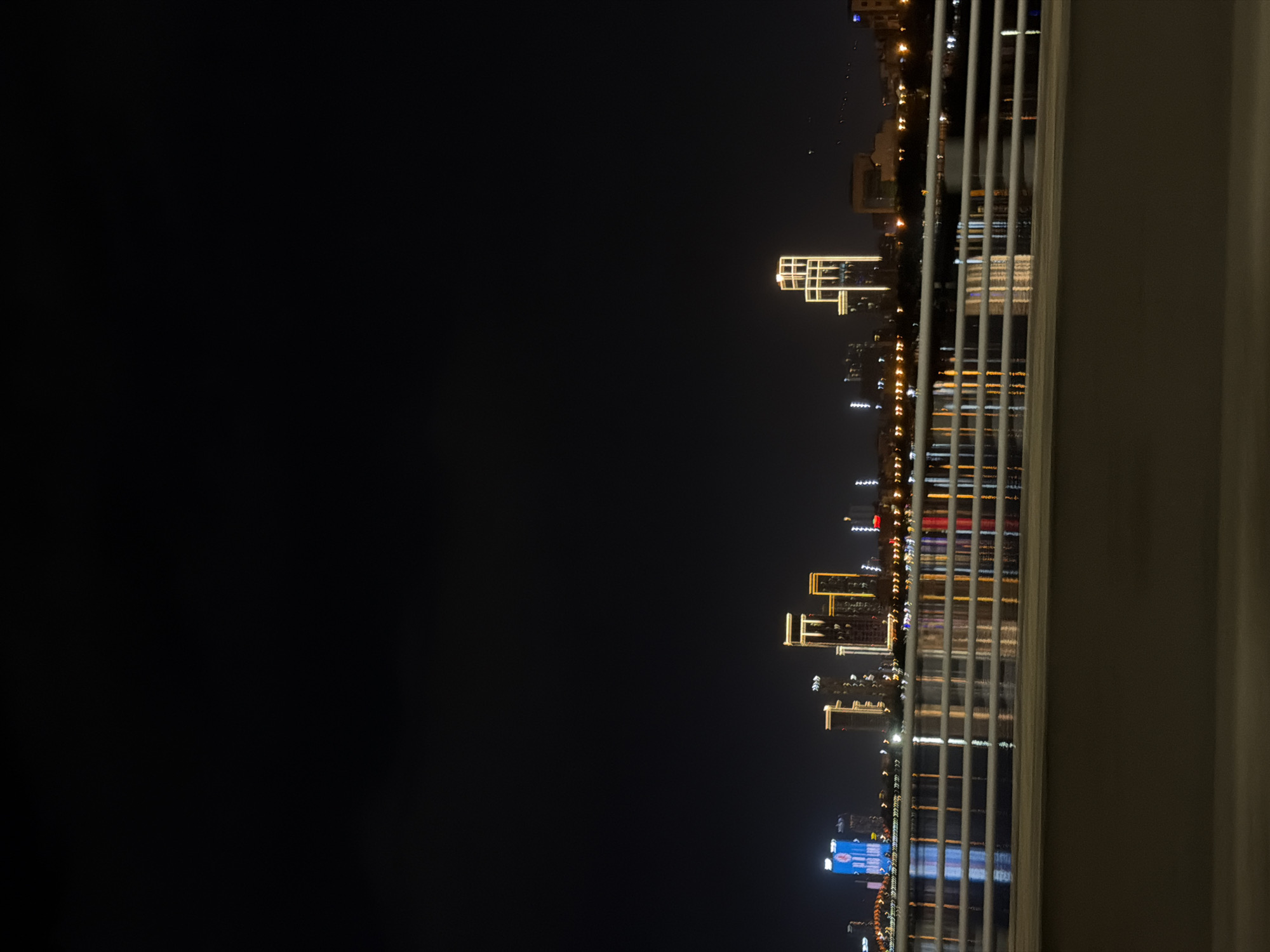 The image displays a nighttime cityscape of Da Nang, Vietnam, as viewed from an elevated position, possibly a balcony or window. The scene is dominated by the dark expanse of the night sky on the left, contrasting with the illuminated skyline of buildings on the right. The buildings are adorned with various lights, including warm yellow, white, and some blue hues, creating a vibrant and somewhat reflective effect on the water below.
Several tall buildings of varying architectural styles are visible. One prominent building features a unique, multi-layered geometric design illuminated with bright yellow lights. The lights from the buildings are reflected in the water, creating a shimmering effect that is partially obscured by a series of vertical white railings in the foreground. These railings suggest the vantage point is from a structure with a protective barrier.
The time of day is clearly nighttime, given the dark sky and the artificial illumination of the city. There are no discernible people or specific activities taking place, as the focus is on the architectural landscape and the city lights. The weather appears to be clear, allowing for a sharp view of the skyline. The presence of water and the distinctive building designs are location cues that, combined with the provided context of Da Nang, Vietnam, help identify the scene. The overall emotion conveyed is one of urban beauty and tranquility at night. No visible text is present in the image.