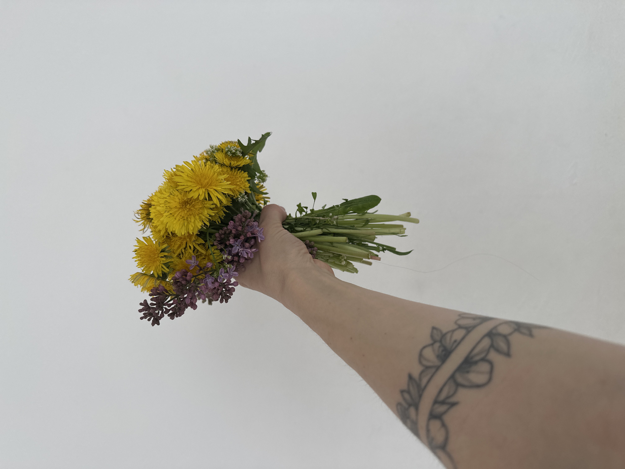 A bouquet of yellow dandelions and purple lilac flowers is being held in a fair-skinned arm with a tattoo of flowers wrapped around the arm, set against a white background. There is no information to suggest this image was taken in Votkinsk, Russia.