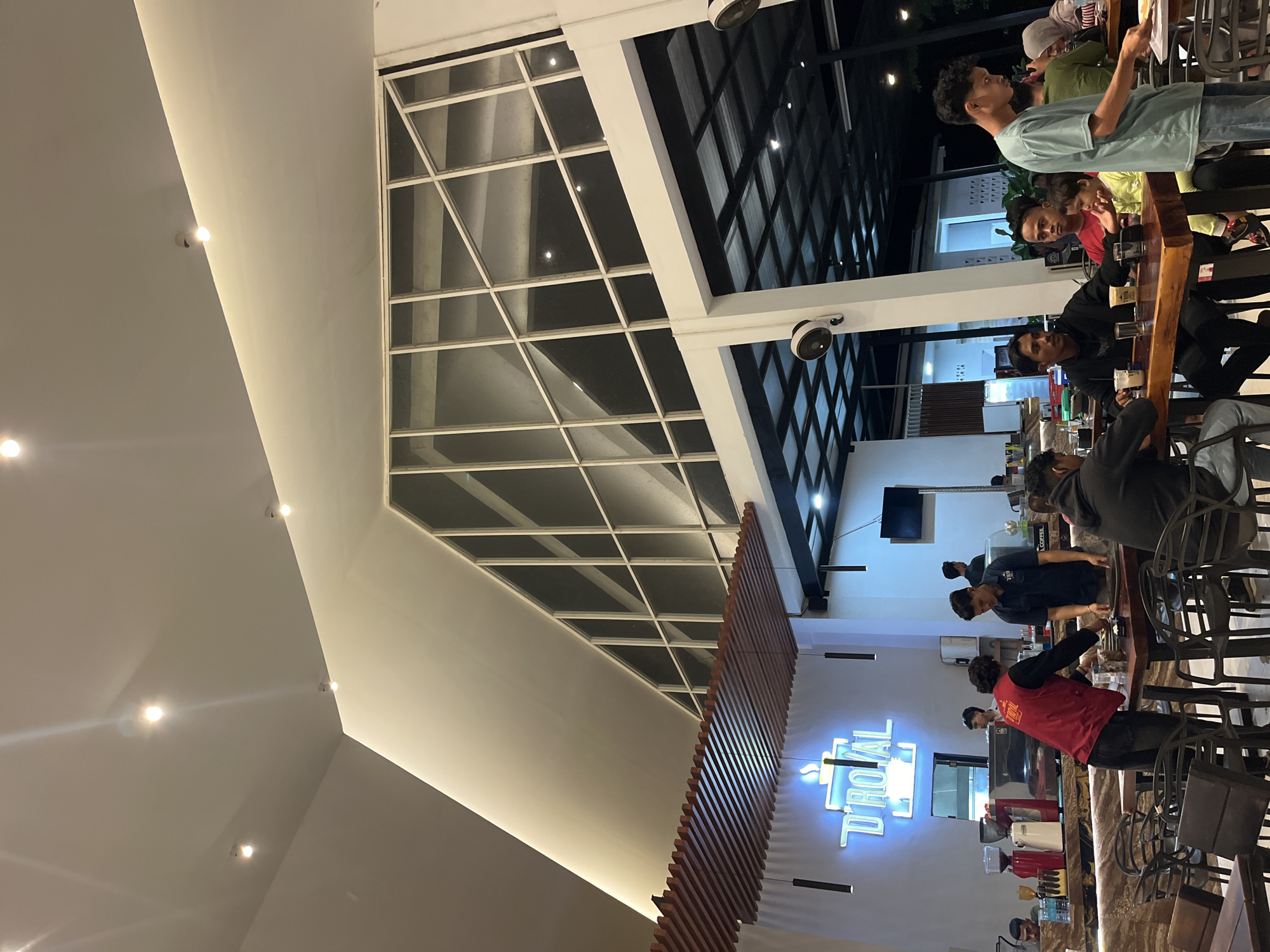 The image depicts the interior of what appears to be a café or restaurant, likely located in Lhokseumawe, Indonesia. Several individuals are present, some seated at a long wooden bar, engaging in conversation and drinking. The café is illuminated with recessed ceiling lights and a neon sign spelling out "D'ROYAL". A prominent architectural feature is a large, geometrically-patterned glass ceiling, partially covered by a wooden slat structure. The overall impression is that of a modern, casual dining or gathering space.