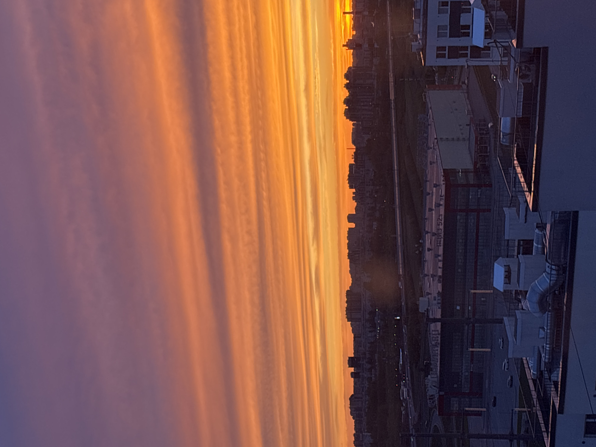 The image shows a sunset over a city. The sky is filled with long, horizontal clouds that are colored in shades of orange and purple. The sun is setting behind the city skyline, casting a warm glow over the buildings. In the foreground, there is a building with various rooftop structures. The location is Unknown, Unknown.
