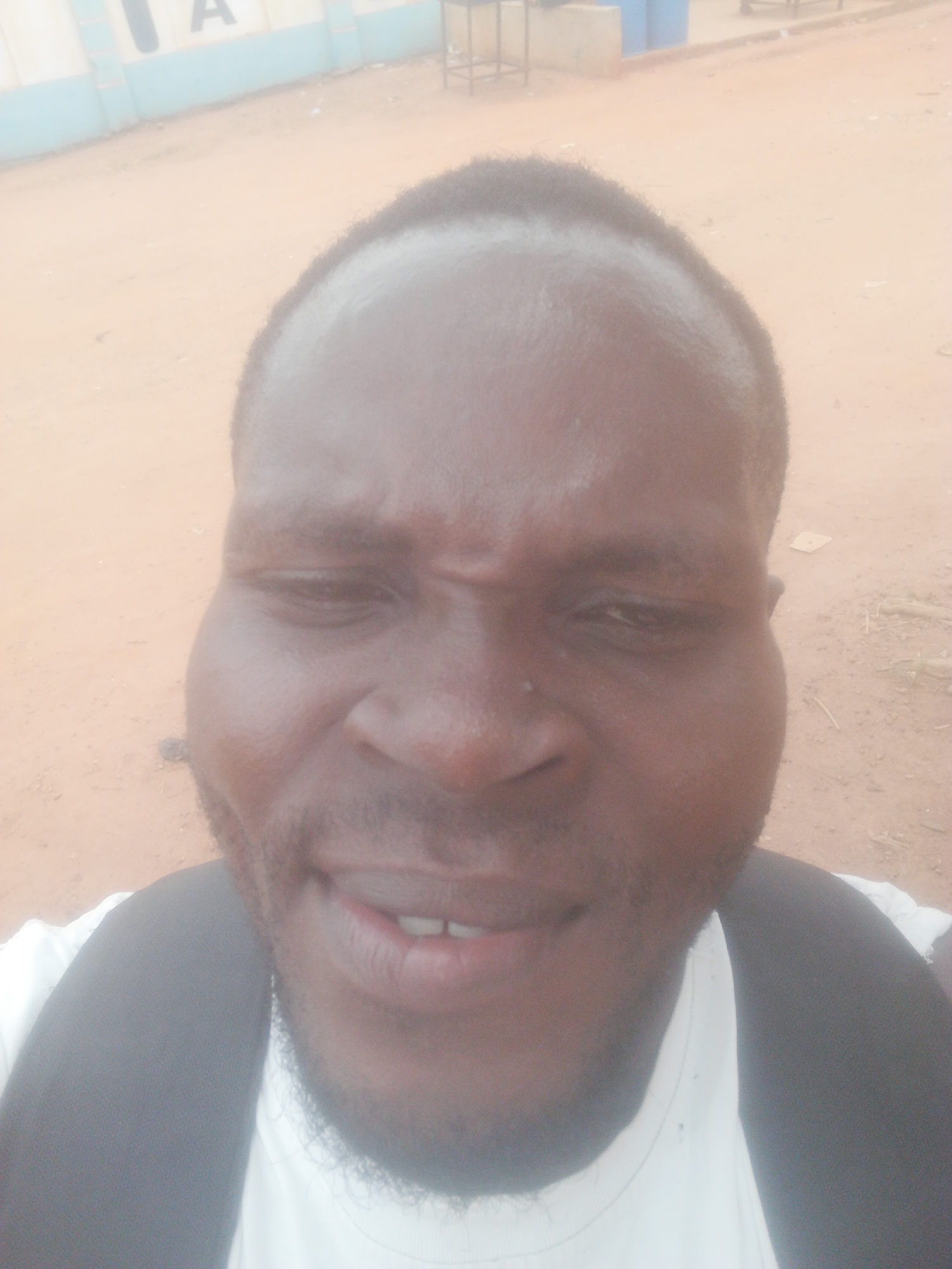 The image is a close-up selfie of a dark-skinned man with short black hair and a beard. He is wearing a white t-shirt and a black backpack. His expression is a slight smile or smirk. The background appears to be an outdoor setting with a sandy or dusty ground. A building with blue and white paint is partially visible in the upper left corner. Based on the prompt information, this image was taken in Ikorodu, Nigeria.