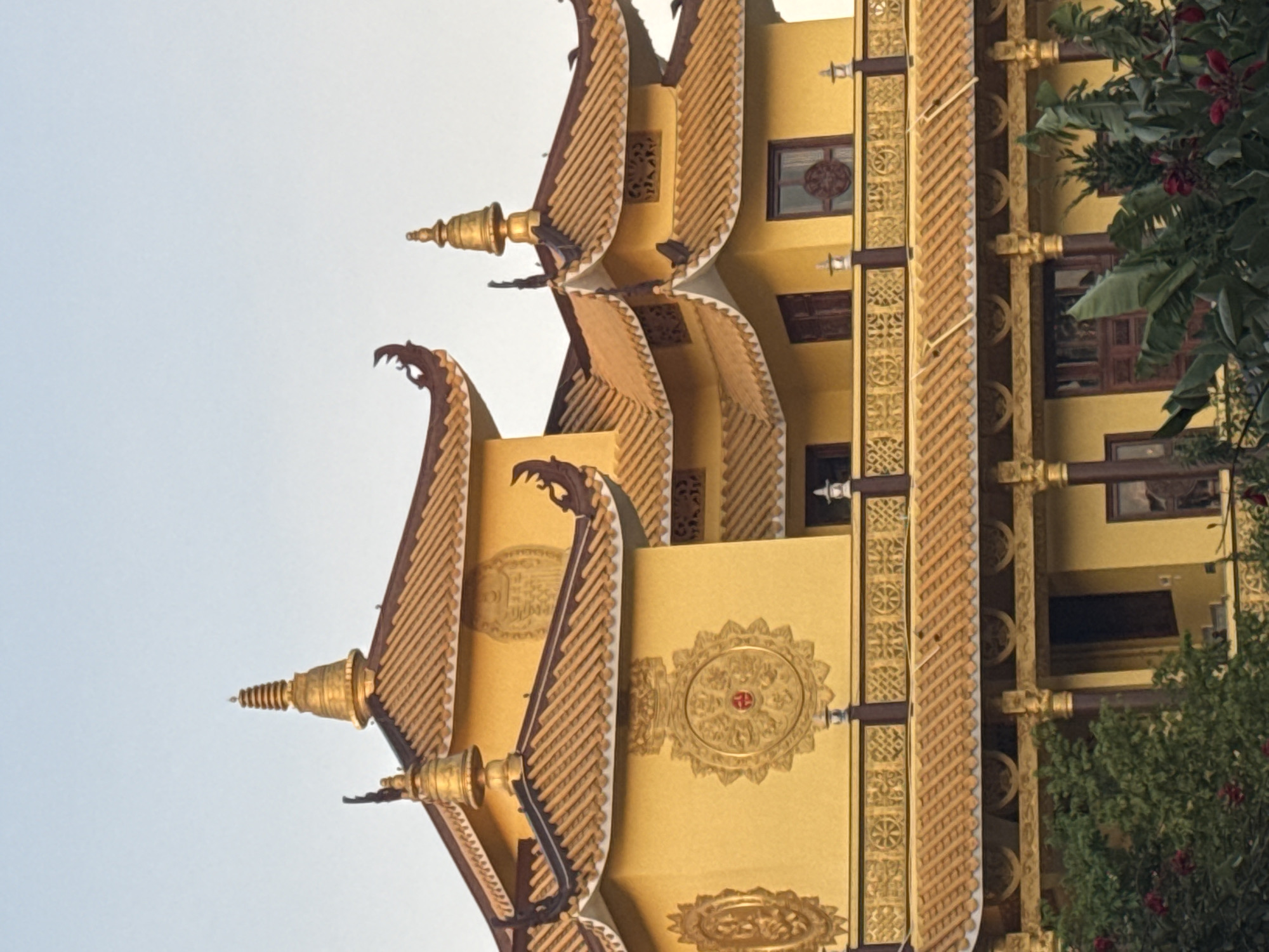The image shows the exterior of a yellow Buddhist temple with ornate gold-colored roofs and decorative elements. The temple features multiple tiers of curved, tiled roofs, topped with golden stupas and finials. Intricate golden circular medallions adorn the walls, some containing red symbolic designs. A decorative railing with repeating patterns runs along the upper level. Lush green foliage, including large leaves and some red flowers, partially obscures the lower portion of the building. The sky above is a pale, clear blue, suggesting it might be morning or late afternoon. This is a view of the Truc Lam Zen Monastery in Da Lat, Vietnam.