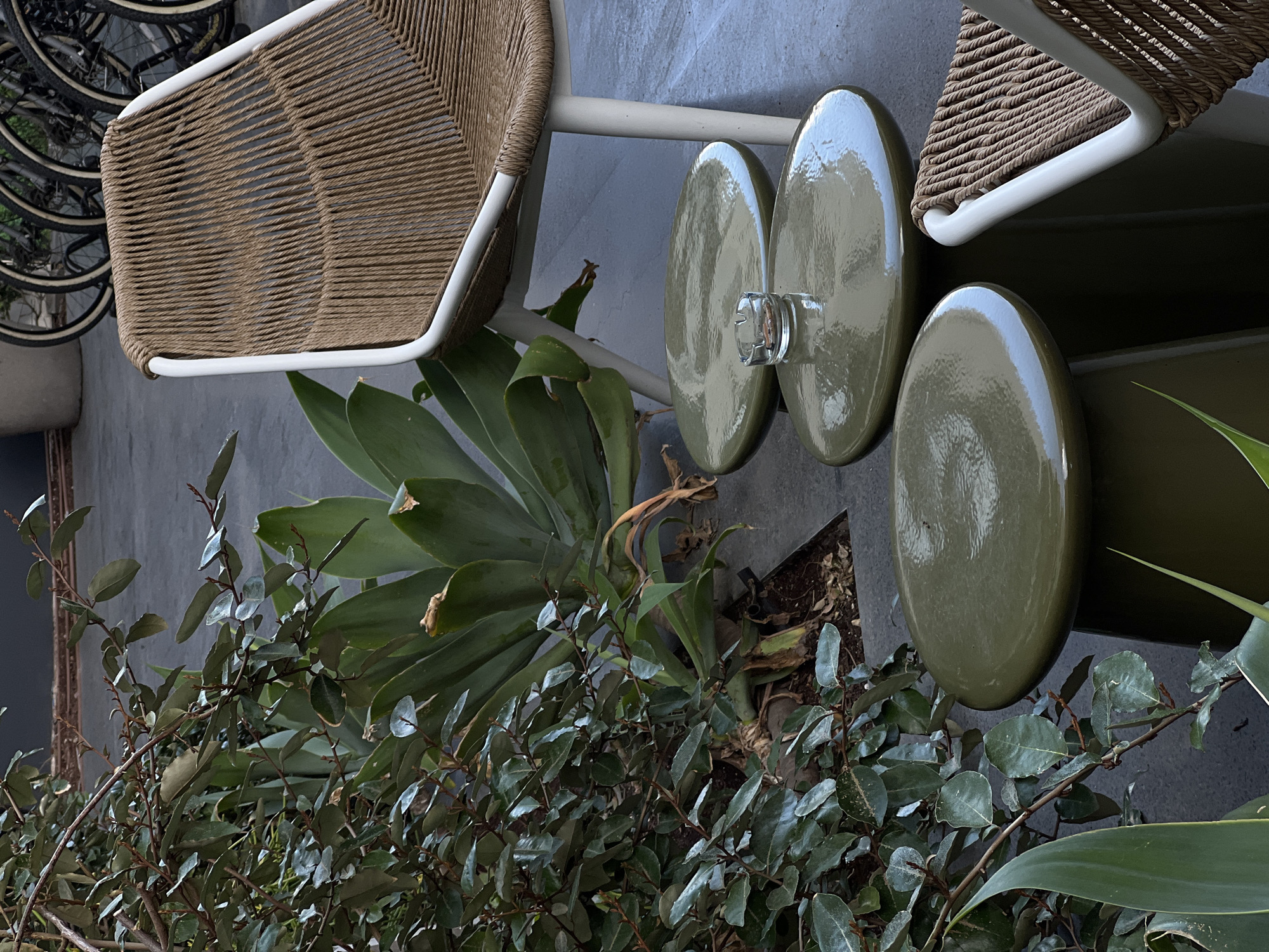 The scene captures an outdoor setting, likely a cafe or patio, featuring a pair of woven chairs with white frames. The chairs are positioned on a gray, paved surface, and lush greenery surrounds them, suggesting a well-maintained urban landscape. Two olive-green, elongated planters with a reflective surface are partially visible, positioned adjacent to the chairs. A stack of bicycles is visible in the background. The environment appears to be in Tel-Aviv, Israel.