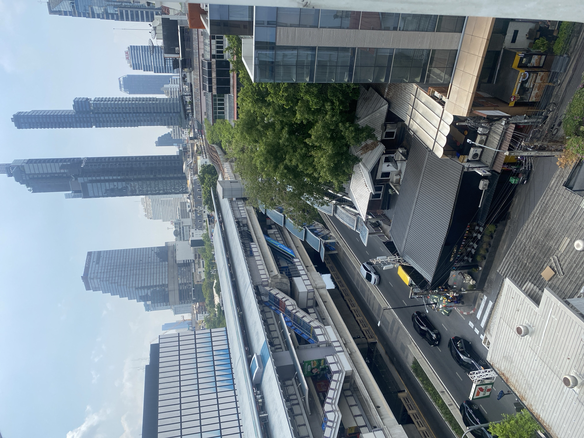 The image shows an elevated view of Bangkok, Thailand. The BTS Skytrain runs parallel to a street with cars and pedestrian crossings. Adjacent to the Skytrain station is a dense urban area with commercial buildings and traditional Thai-style architecture. Tall modern skyscrapers are visible in the background. There are several people in the bottom right.
