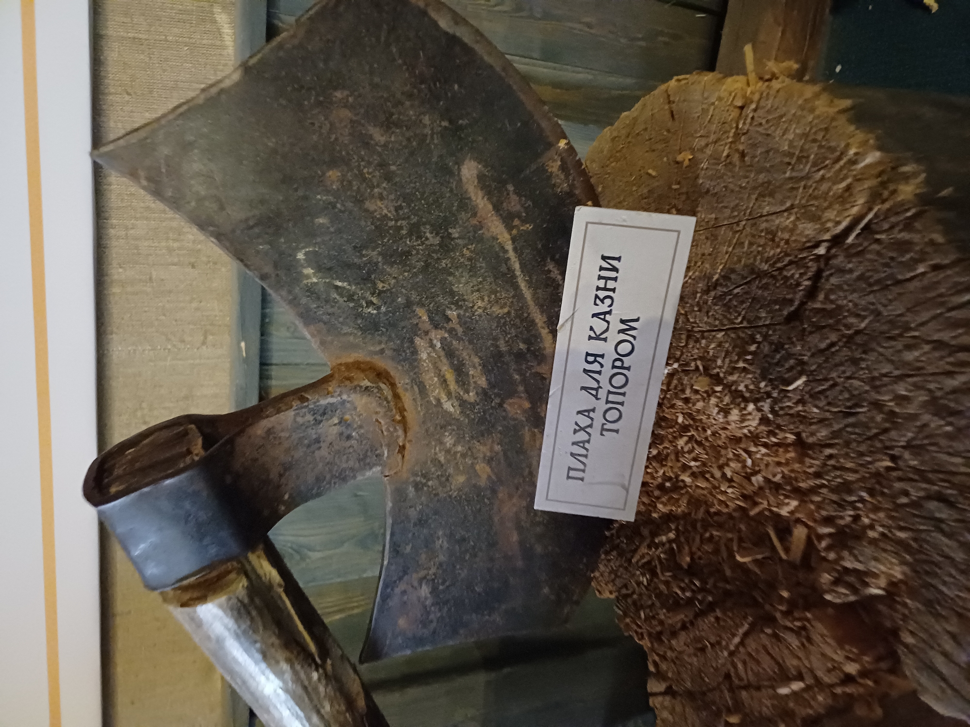 In Estosadok, Russia, an old, rusty axe with a worn wooden handle rests on a thick log covered in wood shavings. A white label is placed on the log, in front of the axe, with Russian text. The background consists of wooden walls.