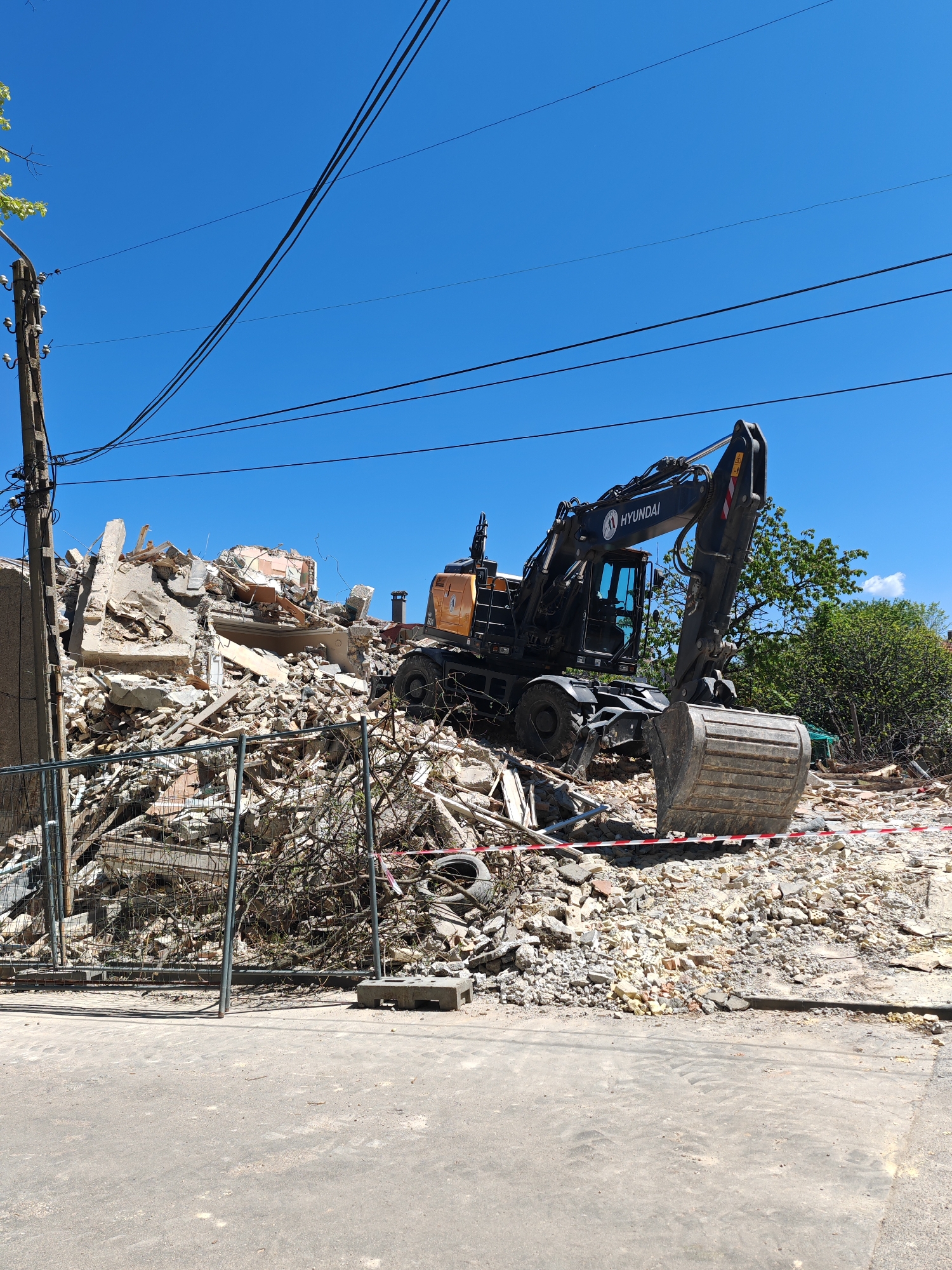 An excavator sits atop a pile of rubble and debris in Shumen, Bulgaria, under a clear, blue sky. The excavator, marked with "HYUNDAI," is black and orange and positioned on top of the demolition remains of a building. The debris consists of concrete, bricks, and metal. A wire fence partially surrounds the area, and red and white tape further delineate the perimeter. An electrical pole stands to the left, with wires extending across the frame. A paved road is visible in the foreground. Trees and bushes are seen in the background.