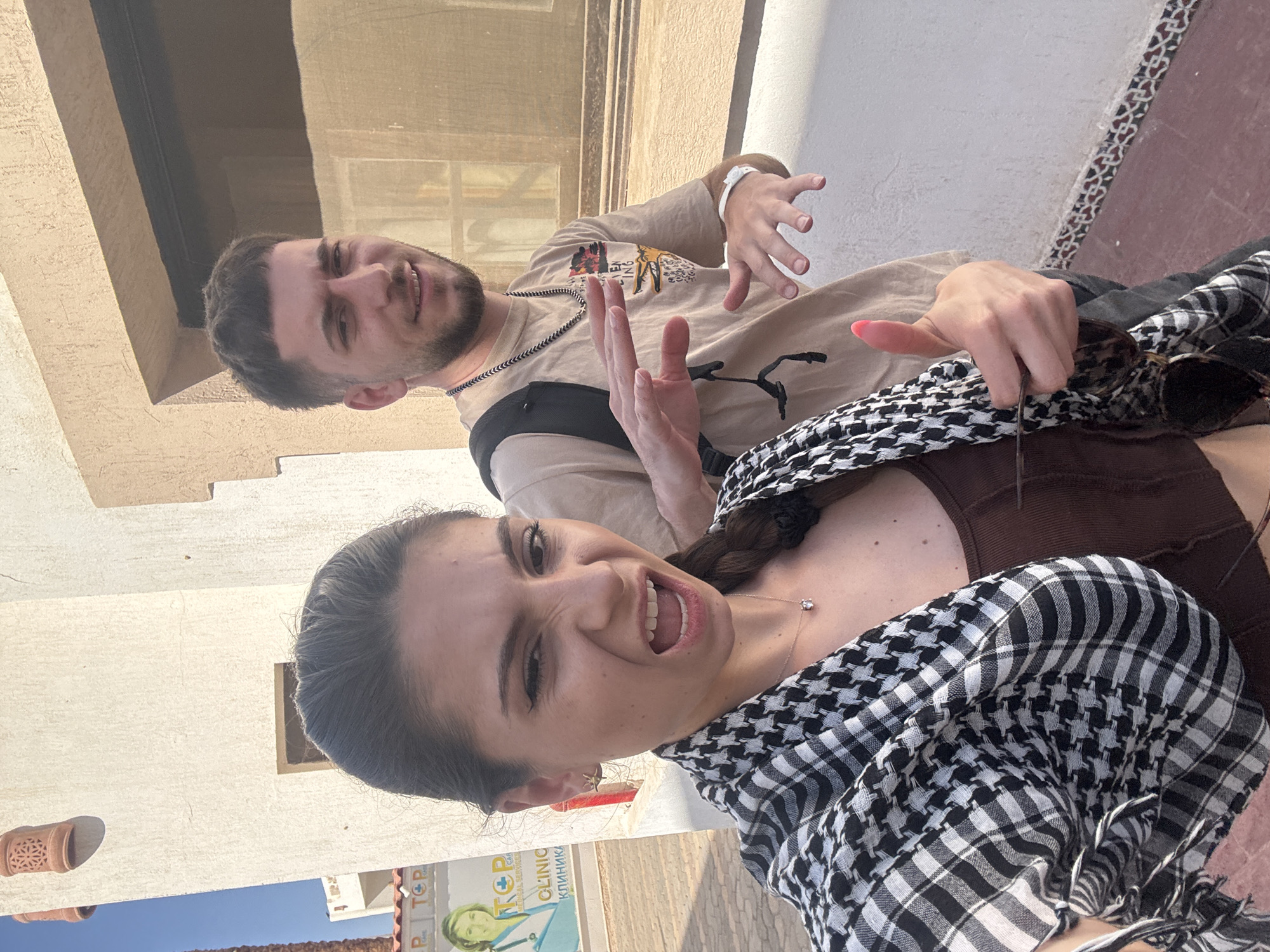 A medium-shot, eye-level photograph features a woman and a man posing for a selfie in an urban setting. The location is in the city of Unknown, Unknown, as per the information provided.
The woman, who is closest to the camera, wears a brown top and a black and white checkered scarf. She has dark hair styled in a braid and a silver necklace. Her facial expression is a playful wink and an open-mouthed smile. The man behind her is wearing a beige t-shirt with a design on it and a black necklace. He is making hand gestures and smiling with an open mouth.
The background includes a building with light-colored walls and a brown window frame. The top part of a sign is visible, with the words "TOP CLINIC" in Cyrillic letters. A blue sky and the tops of other buildings are seen in the distance. The overall setting is bright and sunny.