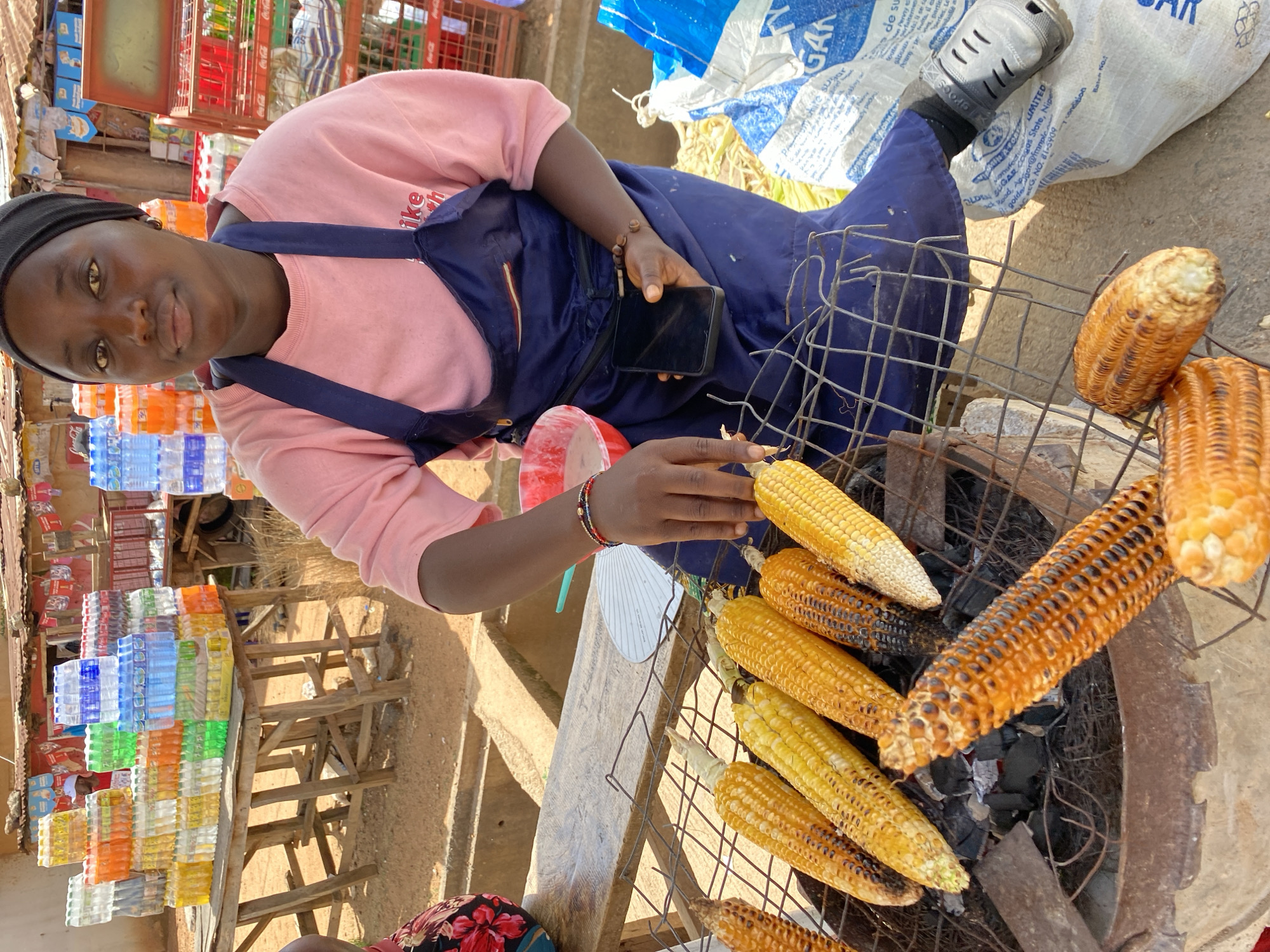 This media file shows a young Black woman in Bukuru, Nigeria, tending to corn being grilled over charcoal. She is wearing a pink sweatshirt and a dark blue apron, with a black head covering. Her left hand, adorned with beaded bracelets, is reaching towards the corn, while her right hand holds a smartphone. The scene is set outdoors, likely at a market stall, as indicated by stacks of bottled water and drinks on a wooden shelf to the left. In the background, there are large bags, one with text including "GAN" and "SUGAR". The corn is arranged on a wire rack over glowing coals, with some pieces showing charring from the grilling process. The lighting suggests it is daytime. The woman's expression is neutral, as she focuses on her task. The presence of the smartphone suggests a blend of traditional commerce with modern technology.