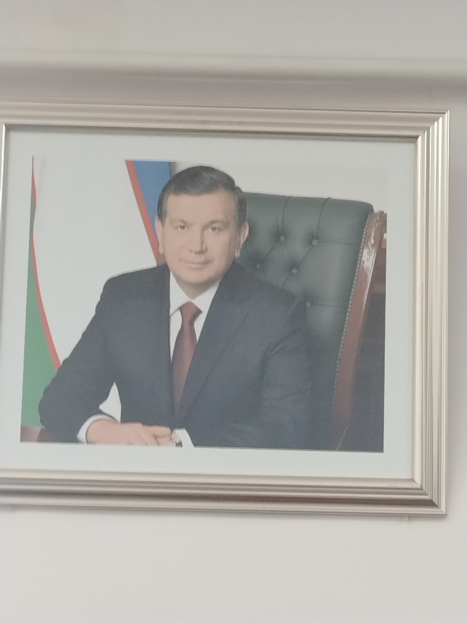 The image is a framed portrait of Shavkat Mirziyoyev, the President of Uzbekistan. He is seated in a dark leather chair, wearing a dark suit, white shirt, and a burgundy tie. He is looking directly at the camera with a slight smile. Behind him, a flag with green, white, and red stripes is visible. The portrait is hung on a white wall, suggesting it is in an office or official setting. The frame is silver and has a ribbed texture. The overall impression is formal and presidential. This portrait could be located in any official building or public space within the city of Nurata, Uzbekistan, as it is a representation of the country's leader.