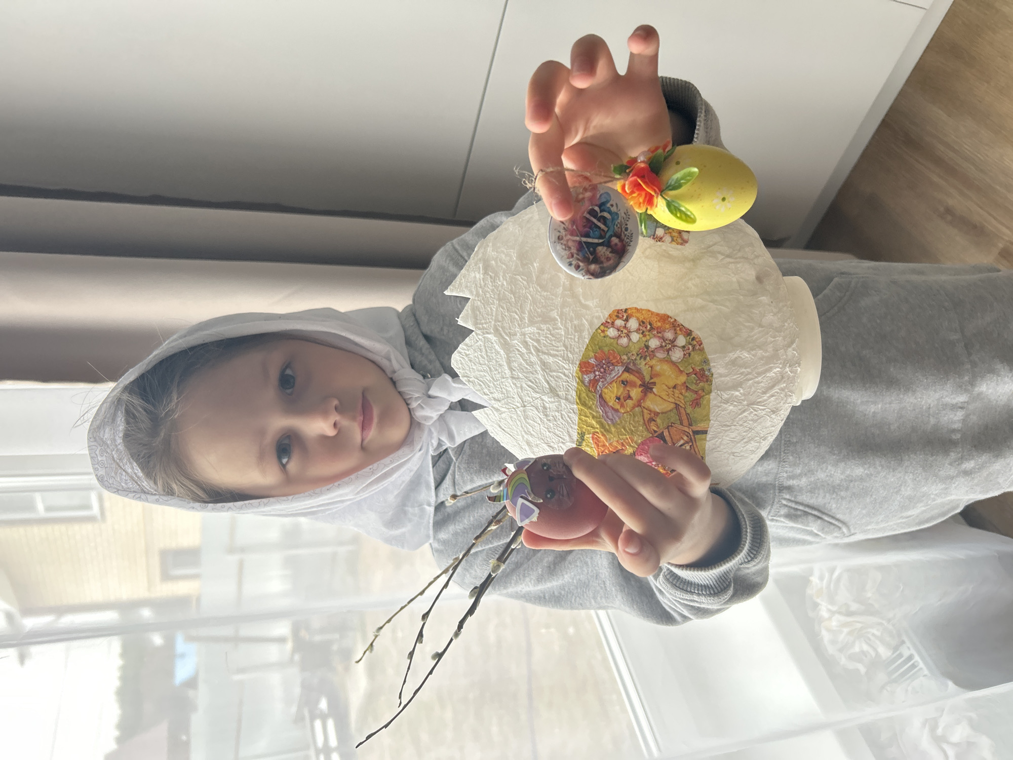A child in a grey hooded sweatshirt and white headscarf stands indoors, holding decorated Easter eggs and a decorative Easter egg container. The child is Caucasian with blue eyes. One egg is decorated with pussy willows and stickers, another with a small floral decoration. The egg container has images of chicks and flowers on its surface. The background shows a window and a blurred view of buildings outside. The location is the city of Chaikovsky, Russia.