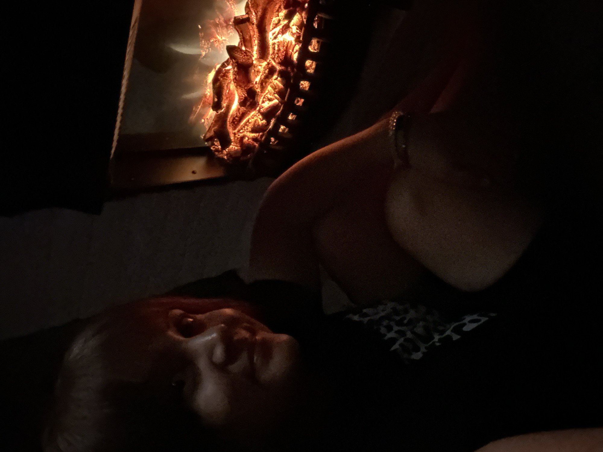 A person is partially visible in the foreground of the image, looking towards an electric fireplace in the background. The person's face is in shadow, but their eyes and mouth are discernible. They are wearing a black shirt with a white pattern. The electric fireplace contains glowing faux logs that emit a warm, orange light, creating a cozy ambiance. The scene is dimly lit, with most of the image in shadow, suggesting it was taken at night or in a dark room. The background is mostly dark, with the fireplace being the primary source of light. The location is described as Unknown, Unknown.