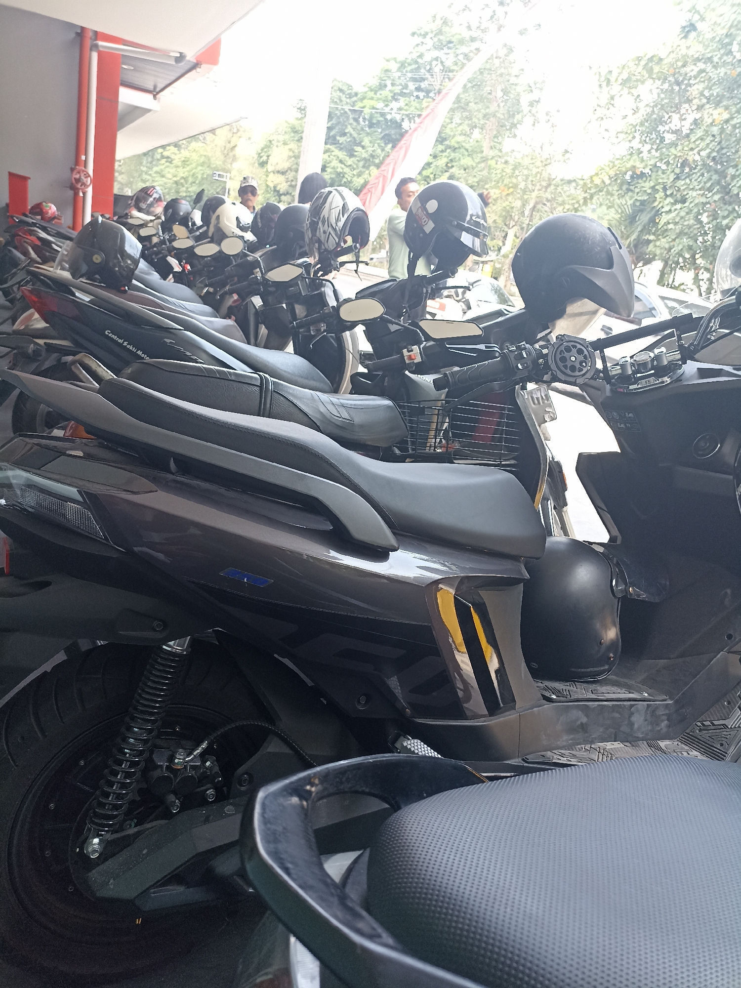 A group of motorcycles are parked in what appears to be an outdoor parking area, possibly at a dealership or a public space. Several helmets are placed on the seats and handlebars of the motorcycles. In the background, there are trees and a building with a red and white facade. A few individuals are visible in the background, one wearing a light-colored shirt and a cap, and another in a dark shirt. The scene is captured in daylight. The presence of motorcycles and scooters, along with the text "Central Seksi Moto" on one of the vehicles, suggests that this location could be in Surakarta, Indonesia, a city known for its vibrant motorcycle culture and numerous dealerships.