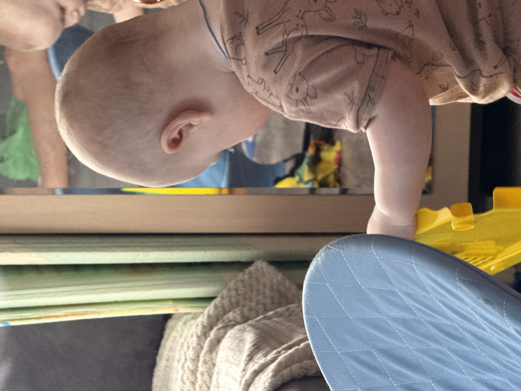 A baby in a patterned shirt is reaching towards a yellow toy near a blue quilted seat. In the reflection of a surface above, a person's face is partially visible. Below, there are stacked objects and a beige knitted item on a gray surface. The location is Кипчак, Russia.