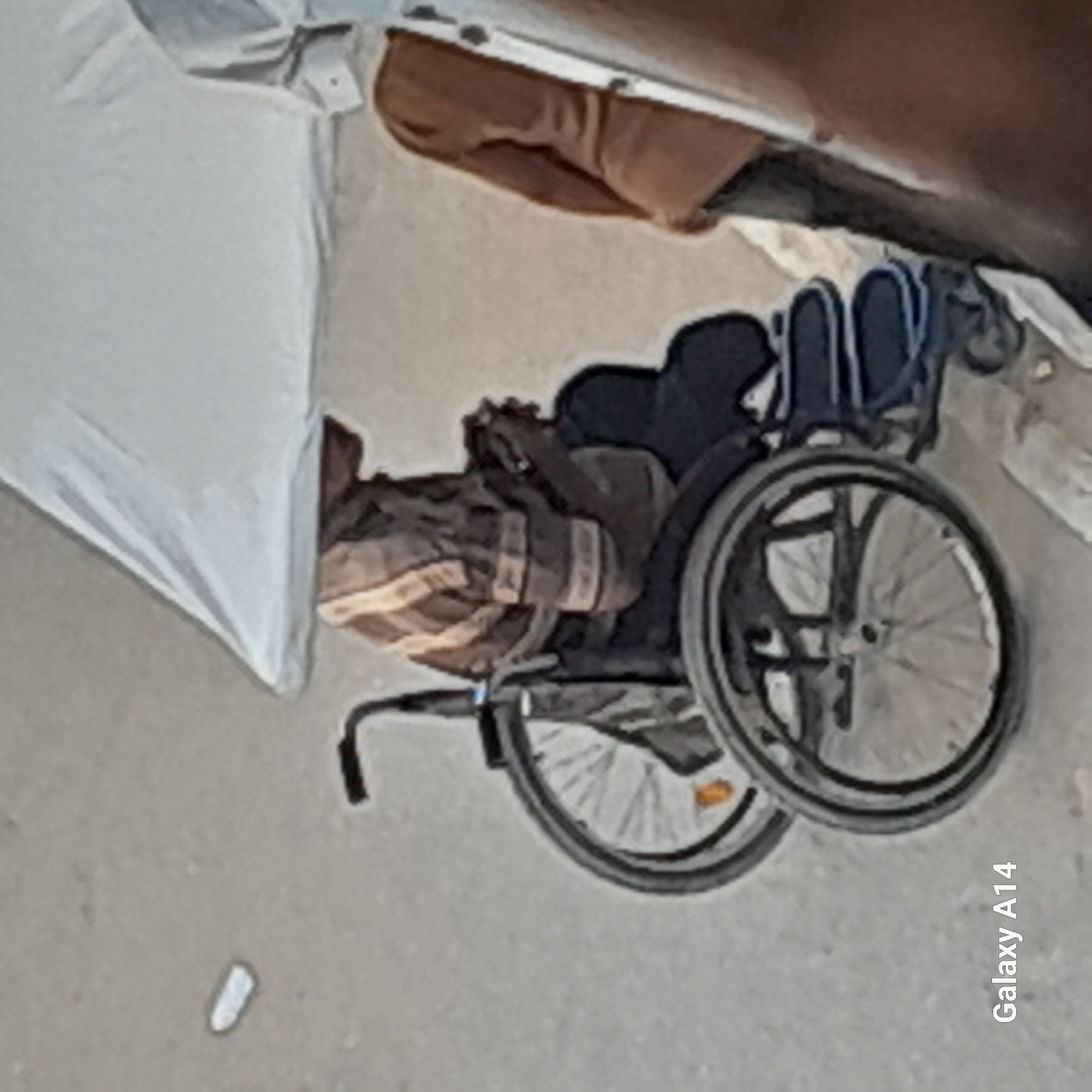 In Khan Yunis, Palestinian Territories, an individual sits in a wheelchair, partially obscured by a light-colored canopy or tent. The person is wearing a horizontally striped long-sleeved shirt and dark pants. Their hands are clasped together. The wheelchair has large spoked wheels. To the right, another person, wearing a brown garment, is visible from the side, also partially under the canopy. The ground appears to be a dusty or sandy surface. A watermark in the lower left corner reads "Galaxy A14".