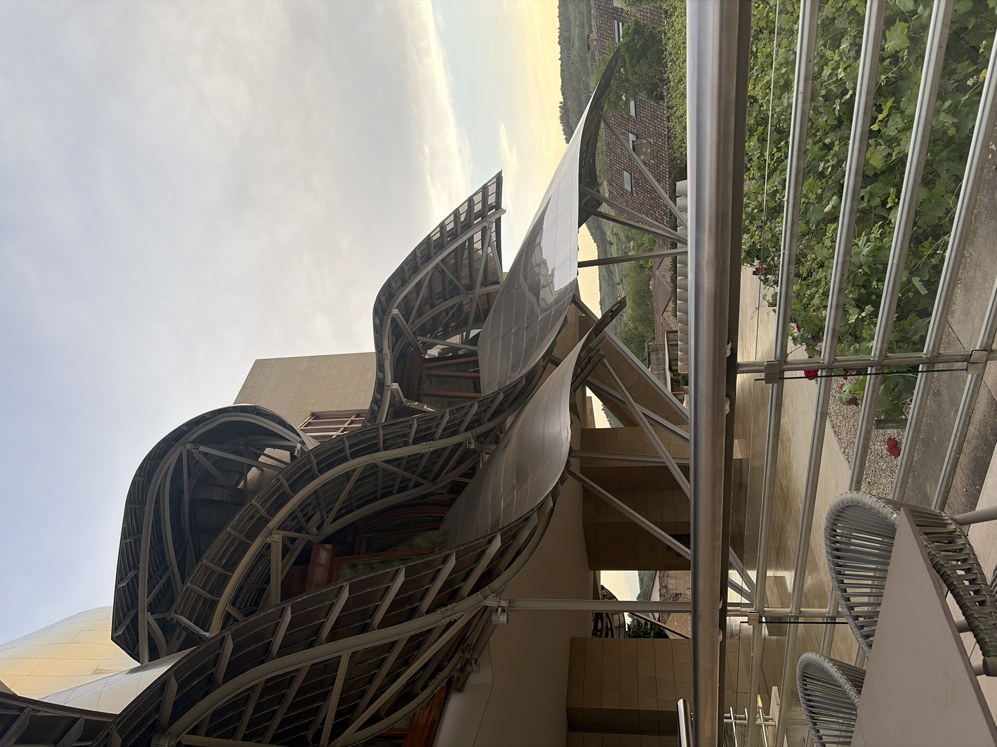 The photo shows a unique structure, possibly part of the Marqués de Riscal winery in Elciego, Spain. It features undulating, metallic ribbon-like designs attached to the building's architecture. The background displays rolling hills with what appears to be vineyards and traditional brick buildings. In the foreground, there's a balcony area with seating, including a table and two chairs. The sky is overcast, suggesting it may be early morning or late afternoon.