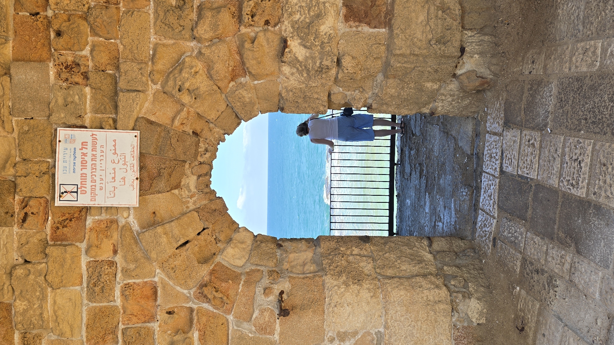 A woman stands looking out at the sea through a stone archway in the city of Acre, Israel. The archway is made of rough-hewn, tan-colored stones. Above the arch, mounted on the stone wall, is a white sign with text and symbols. The sign appears to be a warning or informational notice. Through the arch, a vast expanse of the blue Mediterranean Sea is visible under a bright sky with scattered clouds. The woman is seen from behind, wearing a white tank top and blue striped shorts. She is leaning on a black metal railing that separates the walkway from the sea view. The ground in the foreground is paved with stone slabs. The overall scene depicts a historical and scenic location in Acre.