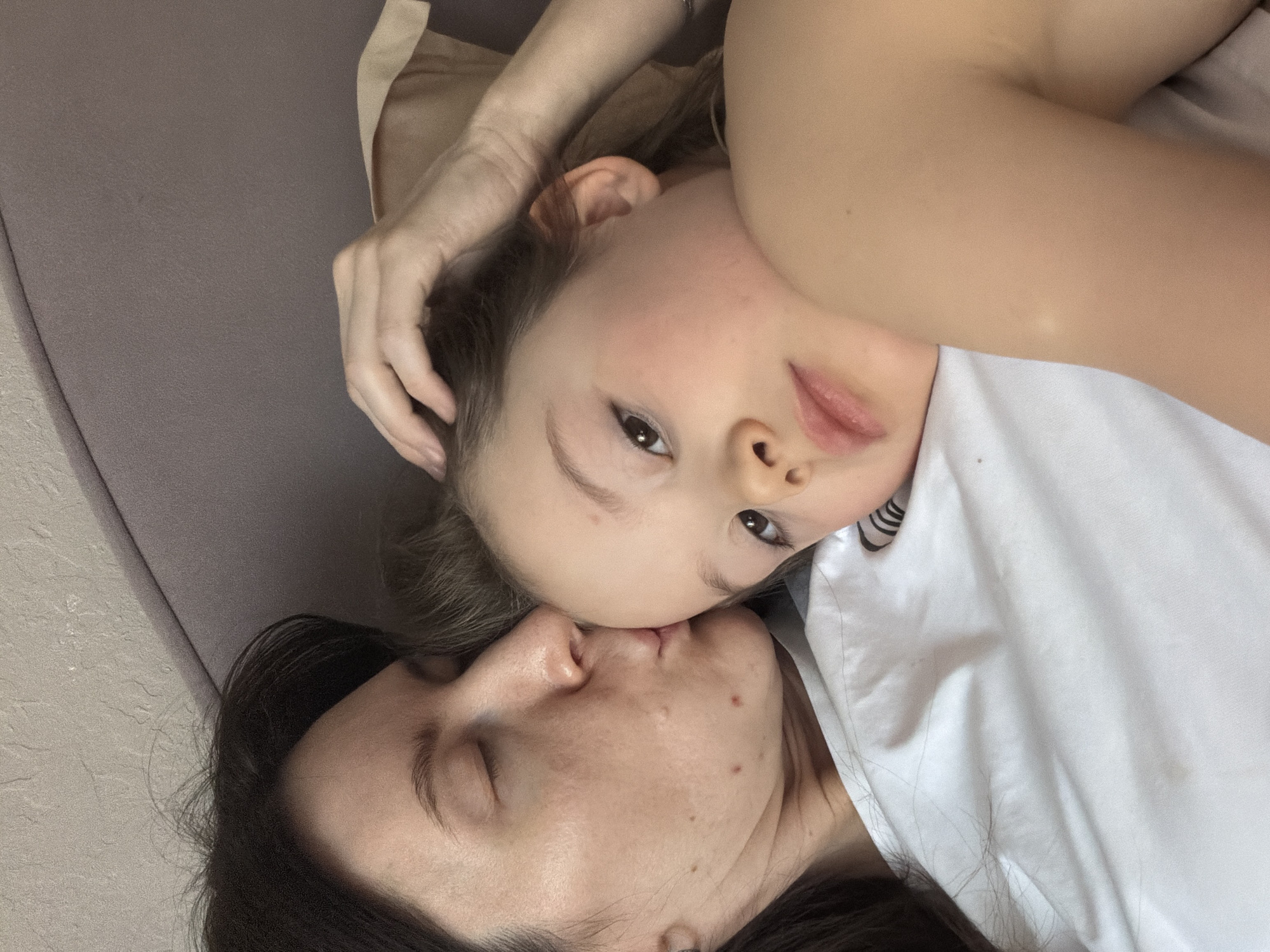 The image displays an adult female and a young child in an indoor setting, identified as Perm, Russia. The adult female, with dark hair, is positioned on the left, with her eyes closed and lips pressed against the child's forehead. She is partially clothed in white fabric. The child, with lighter hair, occupies the right side of the frame, with their right eye open and directed towards the viewer. The child's right hand is raised, with fingers touching their own head, and their right shoulder appears bare. Both individuals are resting on or covered by white bedding. Behind them, an upholstered surface in a greyish-purple hue is visible, likely a headboard, beneath a textured, light-colored wall.