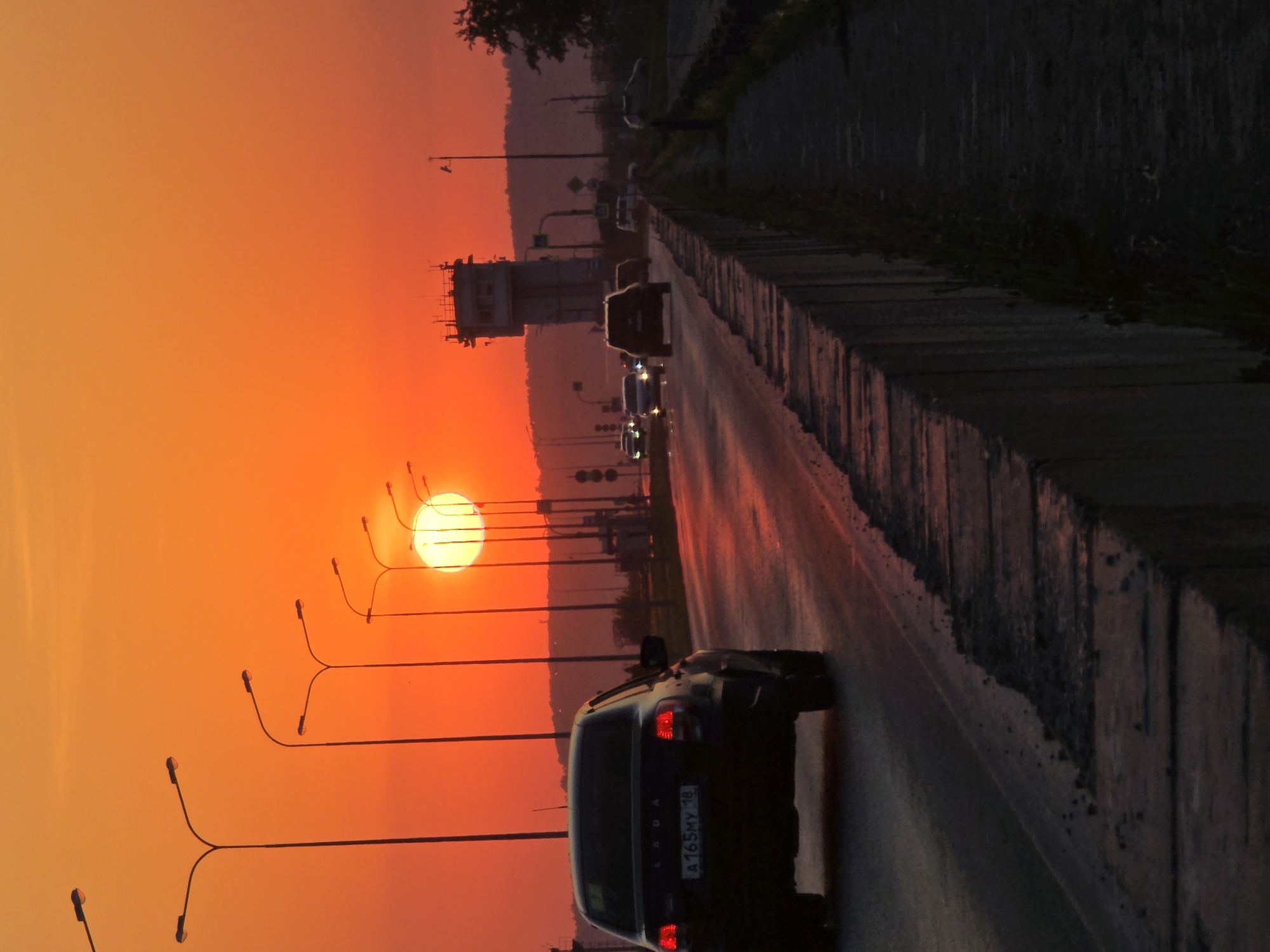 The image captures a sunset over a roadway in Chaikovsky, Russia. Several cars are driving on the wet road, with a black Lada in the foreground. The sky is orange due to the setting sun, which is visible between streetlights. A building is seen along the roadway.