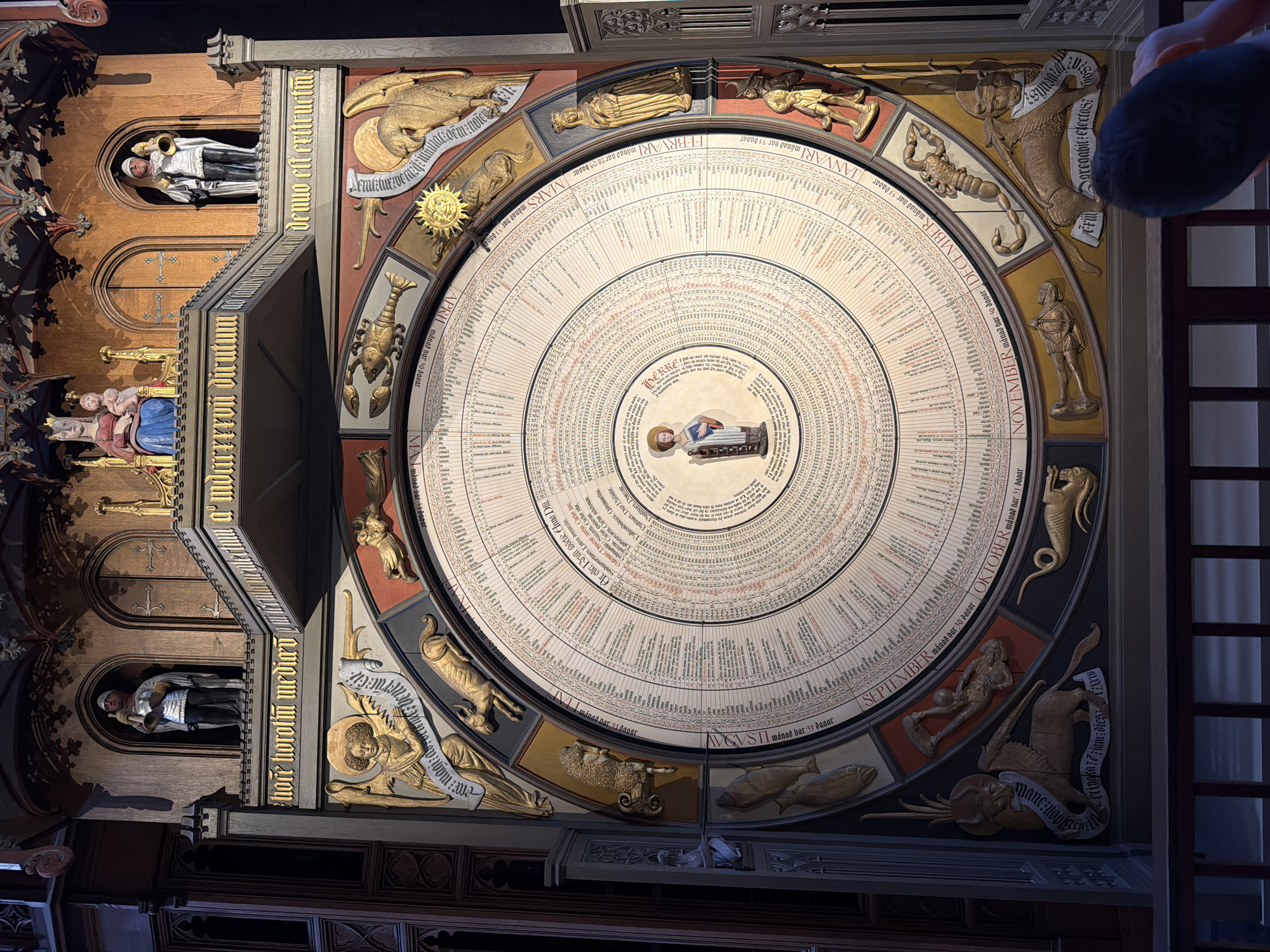 The image shows the astronomical clock in Lund Cathedral, Sweden. The clock is a large, circular mechanism adorned with figures and inscriptions. At the top, above the clock face, are statues of religious figures, including a Madonna and child, and figures in armor holding trumpets. The clock face itself is a complex arrangement of concentric circles with text and numerical data, suggesting it displays astronomical and calendrical information. Around the perimeter of the clock face are depictions of zodiac signs and carved figures representing months or celestial beings, with labels such as "JANUARI," "FEBRUARI," "MARS," "APRILL," "SEPTEMBER," "OKTOBER," and "NOVEMBER." Golden statues of angels and mythological creatures are also integrated into the clock's design. The overall impression is one of intricate craftsmanship and historical significance, characteristic of medieval or early Renaissance astronomical devices. The clock is housed within the dark, ornate interior of Lund Cathedral, with visible wooden architectural elements and decorative carvings.