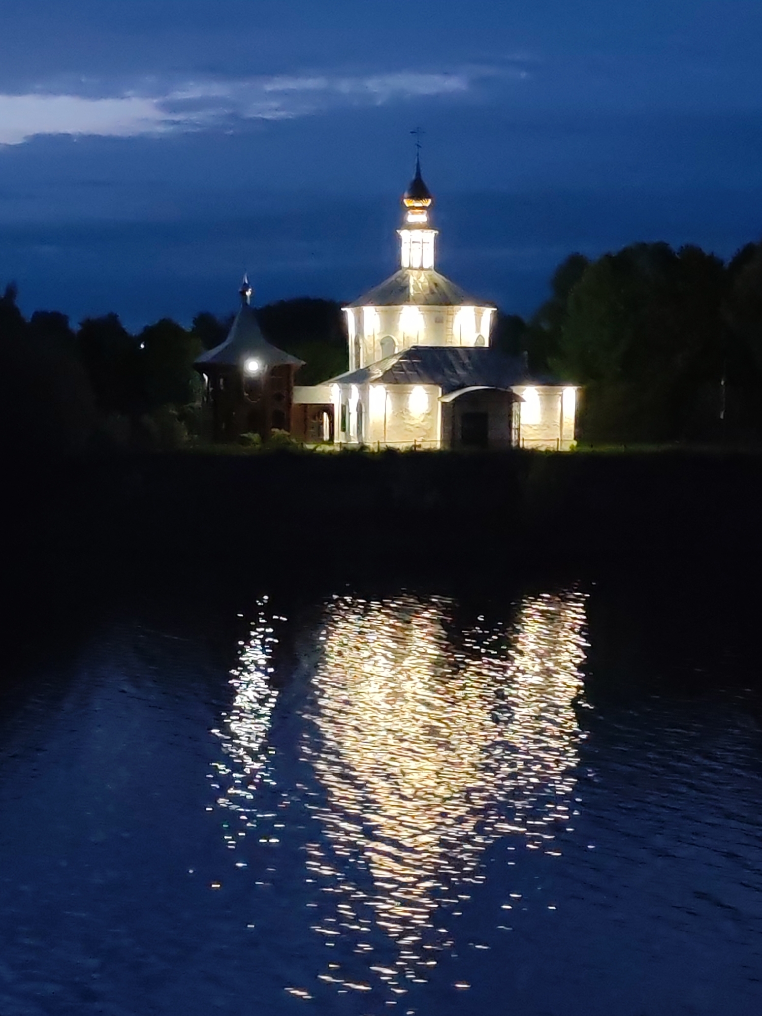 The image depicts the Church of the Praise of the Mother of God in Vasil'ki, Russia, at night. The church is brightly illuminated, and its reflection shimmers on the surface of a body of water, likely a lake or river. The sky is a deep blue, suggesting twilight or a dark evening. Trees line the horizon, silhouetted against the sky. The church's architecture is visible, including its distinct tower and multiple levels.