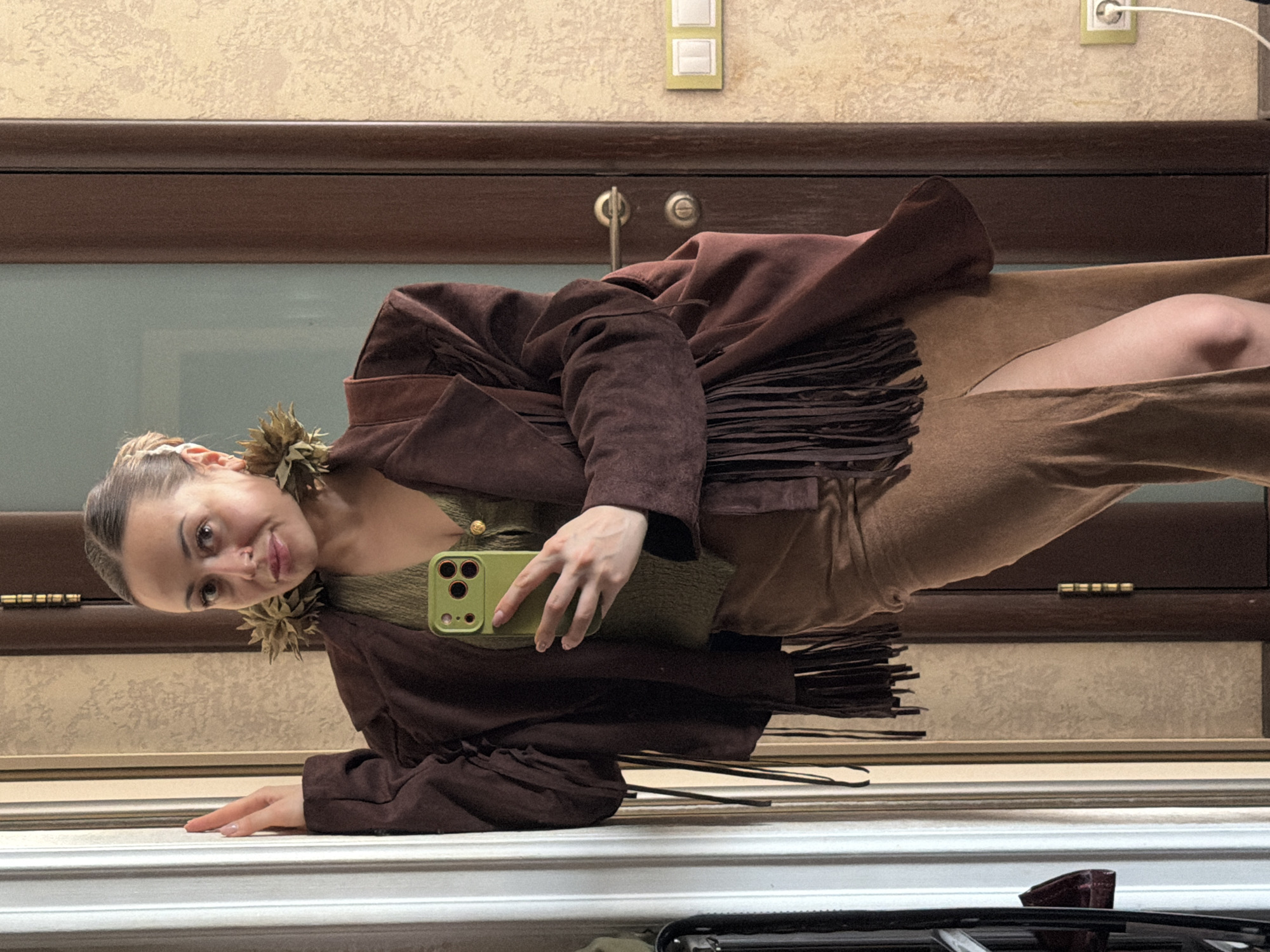A young woman is reflected in a mirror, likely taking a selfie with a green-cased smartphone. She is wearing a brown fringed jacket over a green top, paired with tan pants or a skirt. She has accessories resembling dried flowers on her ears. The background includes a door with dark wood trim, beige wallpaper, and electrical outlets. The location is potentially Krasnoyarsk, Russia.
