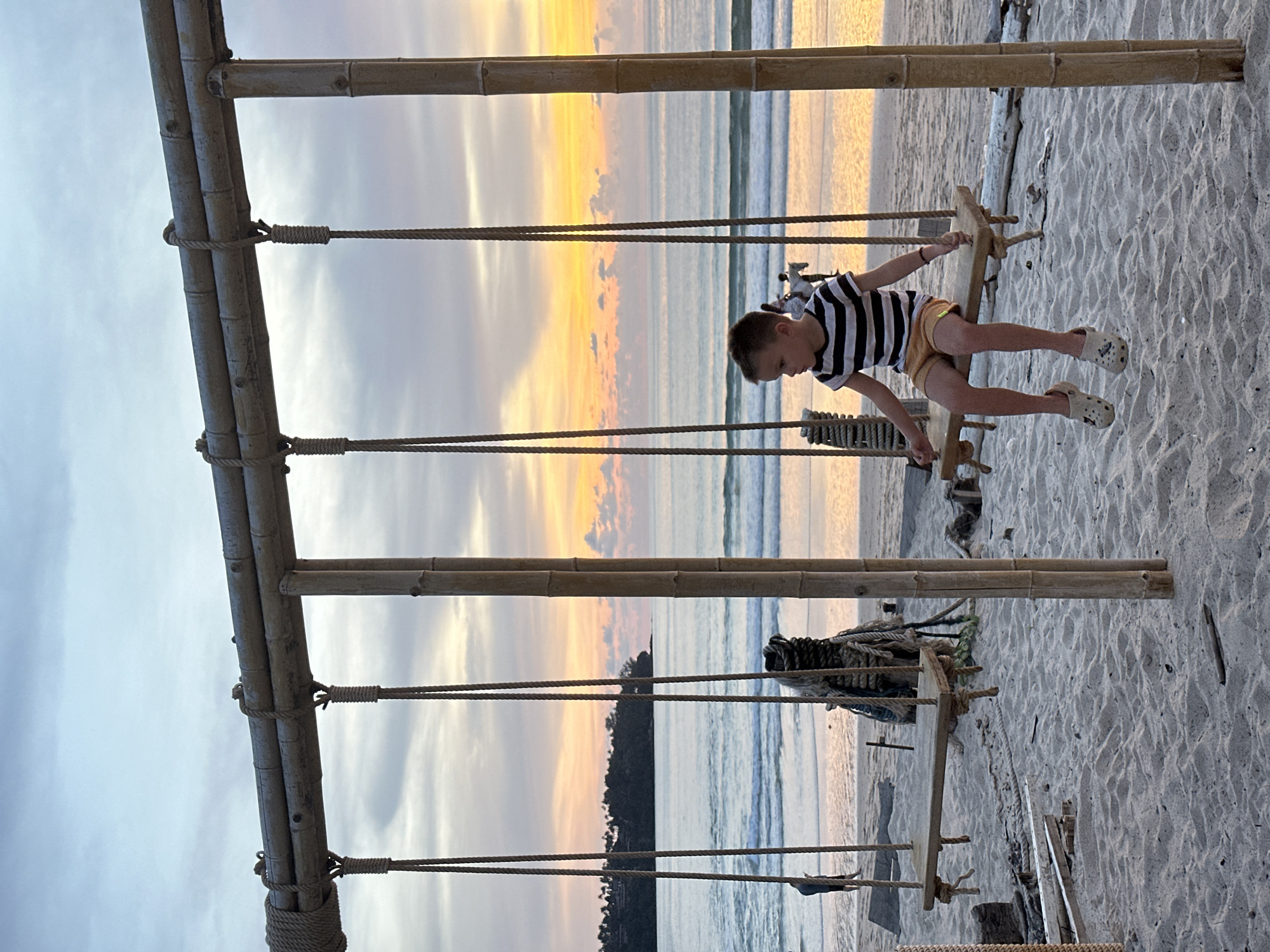 A young boy with light brown hair is sitting on a wooden swing on a sandy beach. He is wearing a black and white striped t-shirt and yellow shorts. The swing is suspended by ropes from a bamboo frame. In the background, the ocean meets the sky at sunset, with the sky displaying orange, pink, and blue hues. Waves are gently rolling onto the shore. A distant island with trees is visible on the horizon. Other swings and a coiled rope structure are also present on the beach. The overall scene is serene and depicts a peaceful moment by the sea.