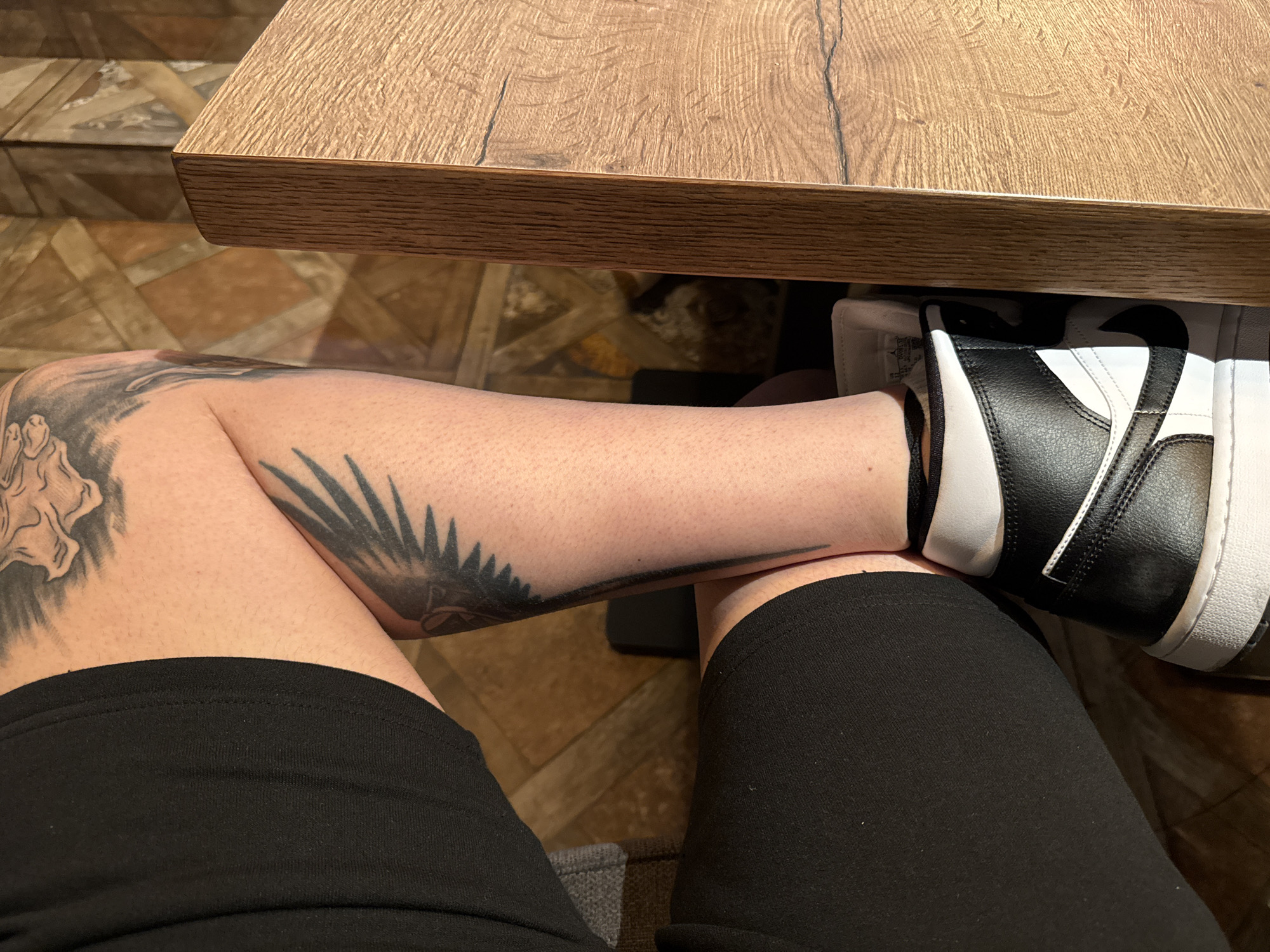 The image shows a close-up view of a person's legs and feet, as well as the underside of a wooden table. The person is wearing black shorts and black and white high-top sneakers. There are tattoos on the person's left leg, including what appears to be a bird or eagle with outstretched wings. The background consists of a tiled floor with a geometric pattern. The scene is indoors, likely in a cafe or restaurant, given the presence of the table and the decor. The specific location in the city of Varna, Bulgaria cannot be determined from the image alone.