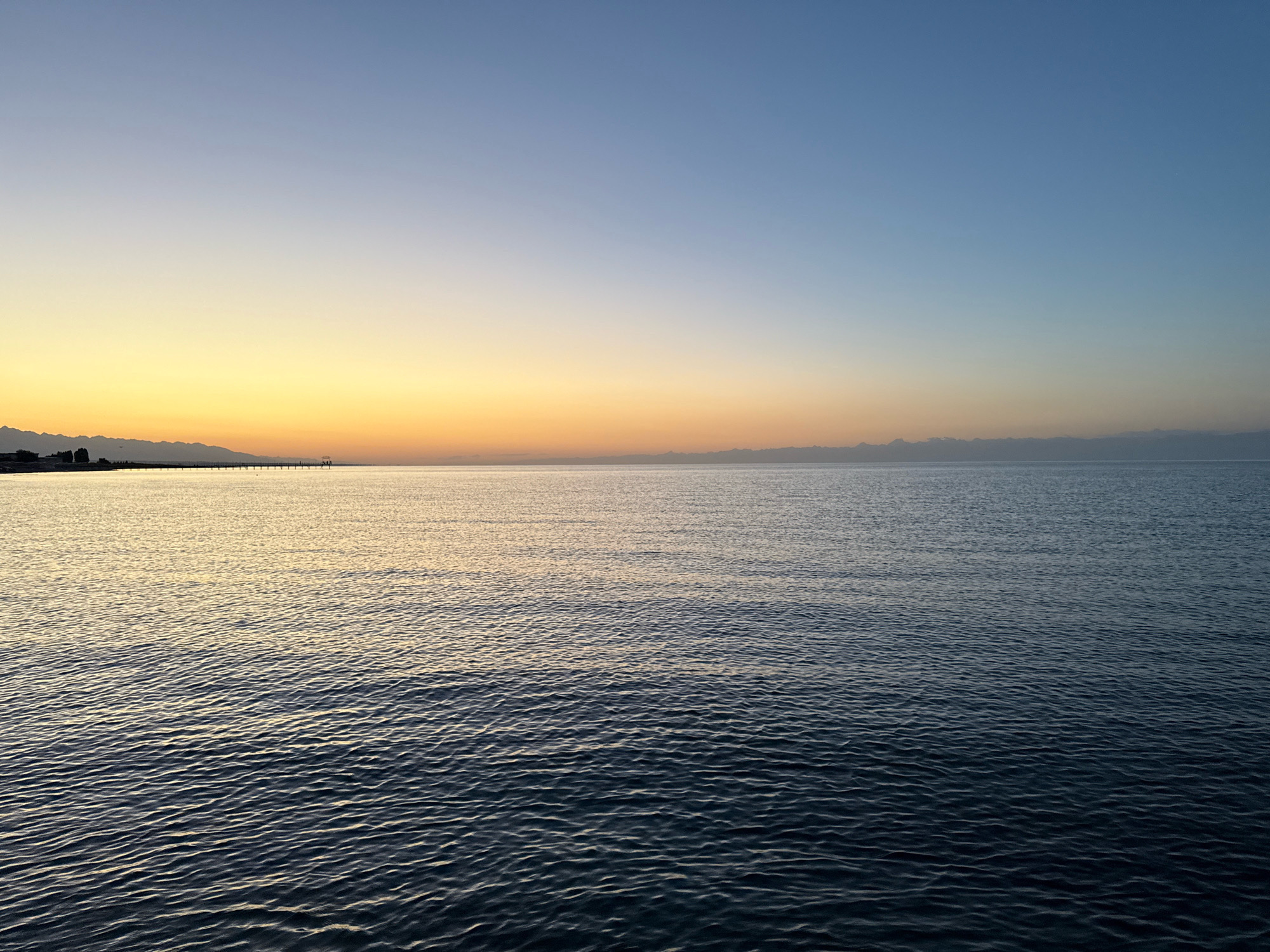 A wide shot captures a tranquil scene at dusk over Issyk-Kul Lake near Bosteri, Kyrgyzstan. The sky transitions from a pale blue at the top to a soft yellow and orange gradient near the horizon, suggesting the sun has recently set or is about to rise. A distant, low mountain range is visible along the far shore, silhouetted against the lighter sky. In the middle ground, a long pier extends into the water, with a few indistinct figures visible on it. The water itself is dark and calm, with subtle ripples reflecting the fading light. The foreground is dominated by the textured surface of the lake, with the ripples becoming more pronounced as they move closer to the viewer. No specific actions or interactions are captured; the image is a static depiction of a serene natural landscape.