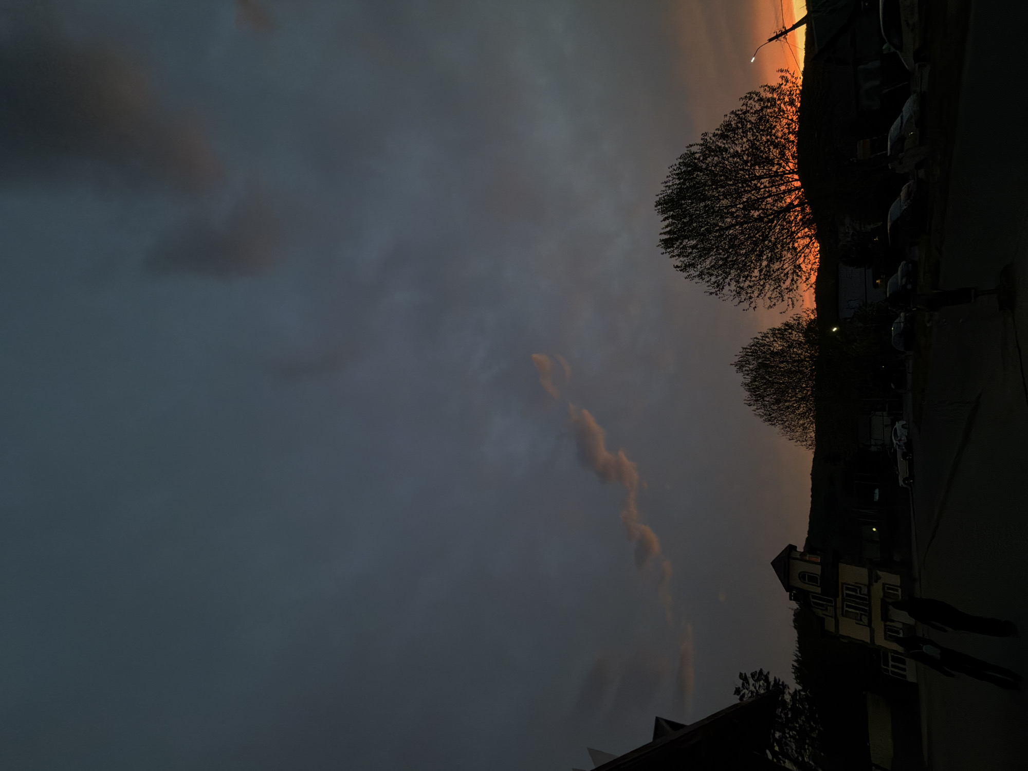 The image captures a dusky scene in Goryachiy Klyuch, Russia. The sky is dominated by a large expanse of dark grey clouds, with hints of orange light near the horizon. Silhouetted trees stand against the glowing sky, and a line of parked cars is visible on the right. A multi-story building with numerous windows is partially visible in the lower right corner. Two shadowy figures appear to be walking on the sidewalk near the building. The scene is dimly lit.