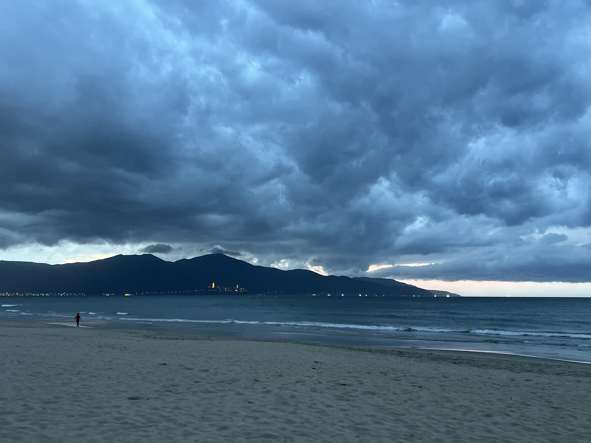 A wide shot of the beach at Đà Nẵng, Vietnam, at dusk showcases a dramatic sky filled with dark, brooding clouds. The choppy turquoise sea reflects the muted light. In the foreground, the sandy beach stretches out, marked by the footprints of a lone figure in a red shirt walking towards the water. In the distance, a dark mountain range forms the horizon, with scattered city lights twinkling along its base and a few boats dotting the sea. The overall mood is somber, hinting at an approaching storm.
