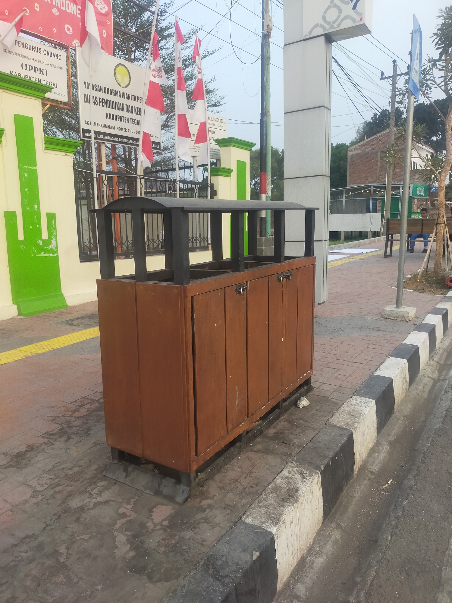 The image shows a brown wooden trash receptacle with a black metal lid frame located on a paved sidewalk in Slawi, Indonesia. The trash can is positioned next to a curb with alternating black and white concrete blocks. In the background, there is a building with green and yellow accents, a street pole with electrical wires, and several Indonesian flags. A person is visible sitting on a bench in the distance. The environment appears to be outdoors, likely in an urban or public area.