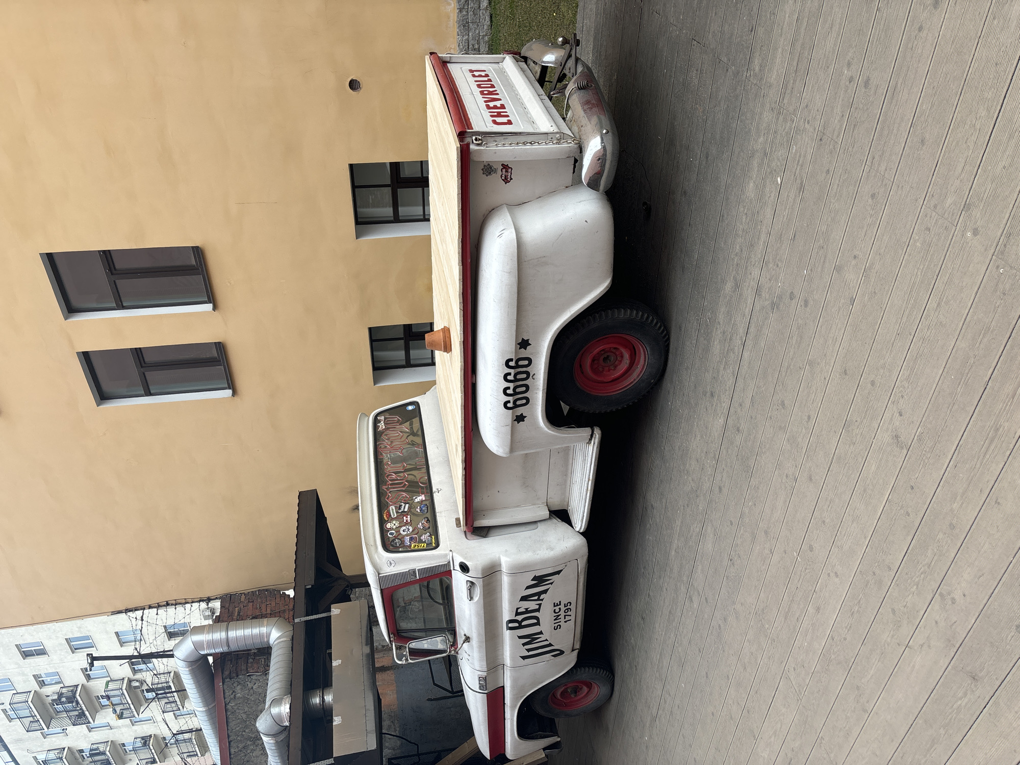 The image depicts a vintage Chevrolet pickup truck parked on a wooden boardwalk in Saint Petersburg, Russia. The truck is painted white with red accents and has the "Jim Beam" logo on its side. The truck's wheel arch has the number "6666". There are stickers on the windshield. The building behind the truck is pale yellow with dark framed windows. A large metal pipe runs along the exterior wall of the building next to the truck. A multi-story building with balconies is visible in the background. The weather appears to be overcast. There are no people visible in the immediate scene.