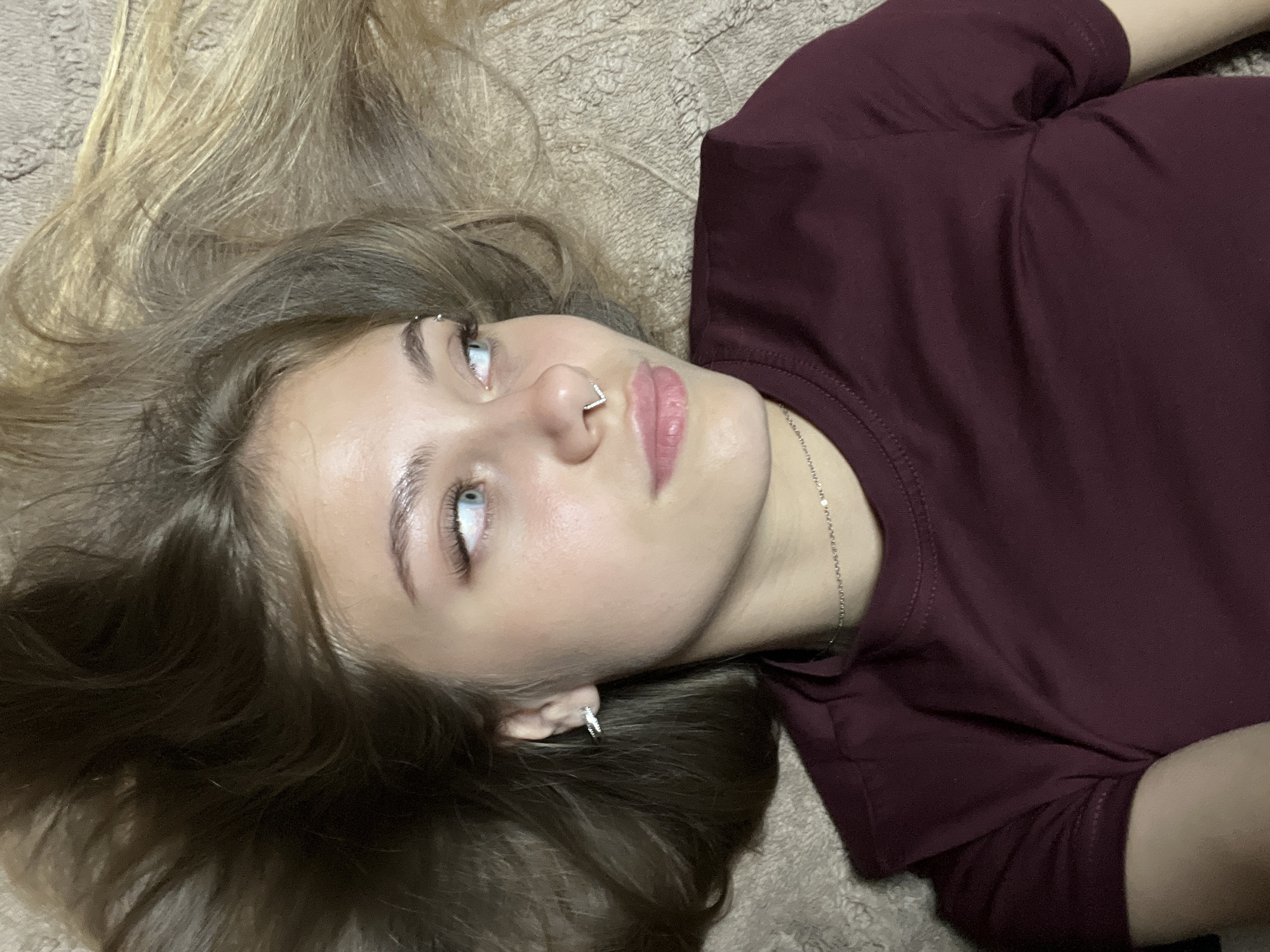 One individual is depicted lying on a light brown, textured surface, appearing to be a soft blanket or carpet. The subject's head is turned to their left, with their gaze directed upwards and slightly to the right of the frame. They have long hair, primarily dark brown with lighter blonde streaks, spread out around their head. The individual has light blue eyes with dark, defined eyebrows and visible eyelashes. A small, V-shaped septum piercing, silver-toned and possibly jeweled, is visible on their nose. Their lips are light pink and slightly parted. A small, silver-toned hoop earring, potentially adorned with small stones, is present in their visible left ear. A thin, silver-toned chain necklace is worn around their neck. The individual is wearing a short-sleeved, dark red or burgundy colored top with a round neckline, showing visible stitching on the shoulders and sleeves. The lighting appears even, suggesting an indoor setting.