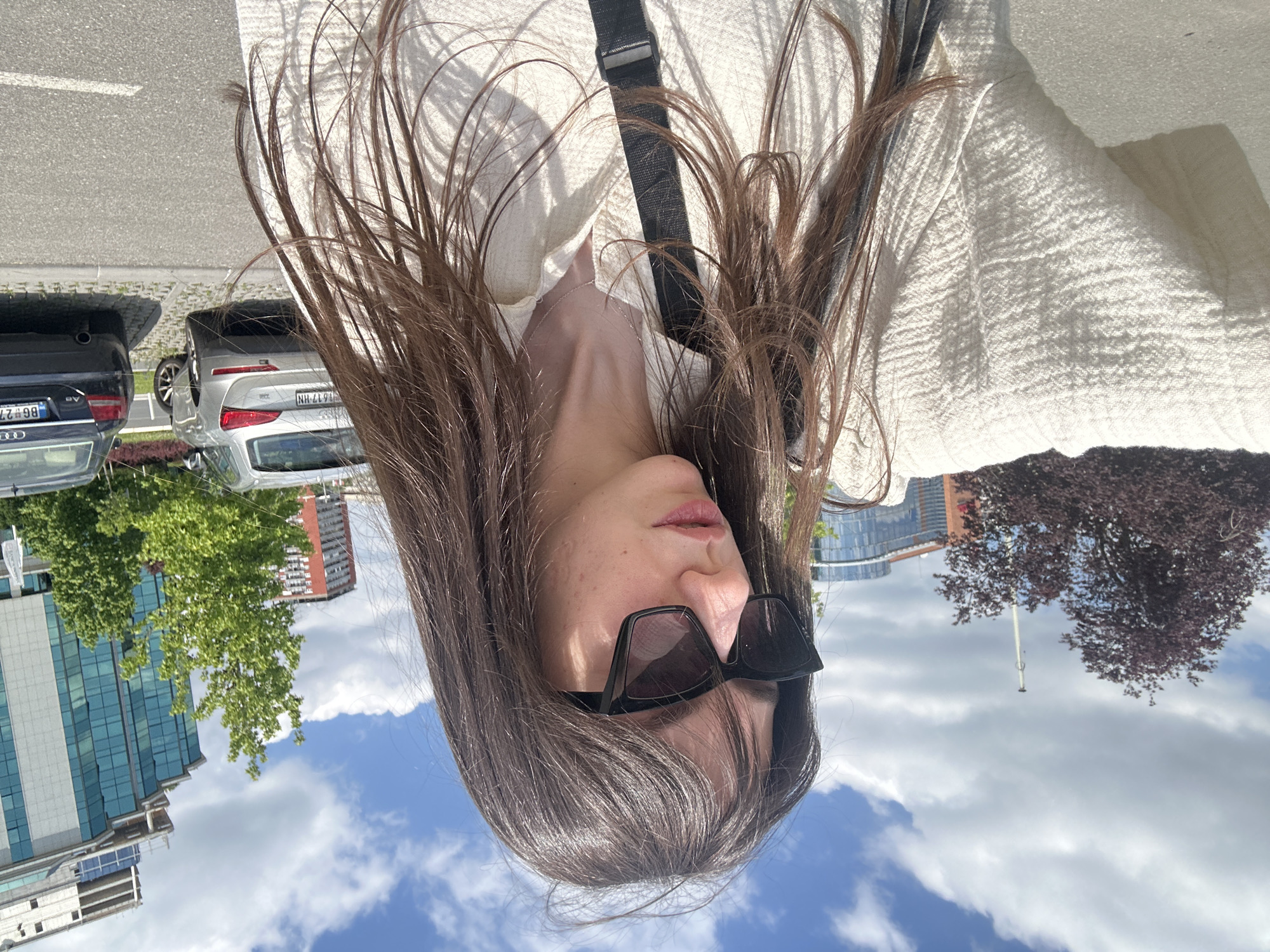 A woman wearing sunglasses and a white shirt is photographed upside down. Buildings and trees are visible in the background, along with cars parked on the side of the road. The cars have license plates from Serbia. The buildings suggest a modern, urban setting. The location is Belgrade, Serbia.