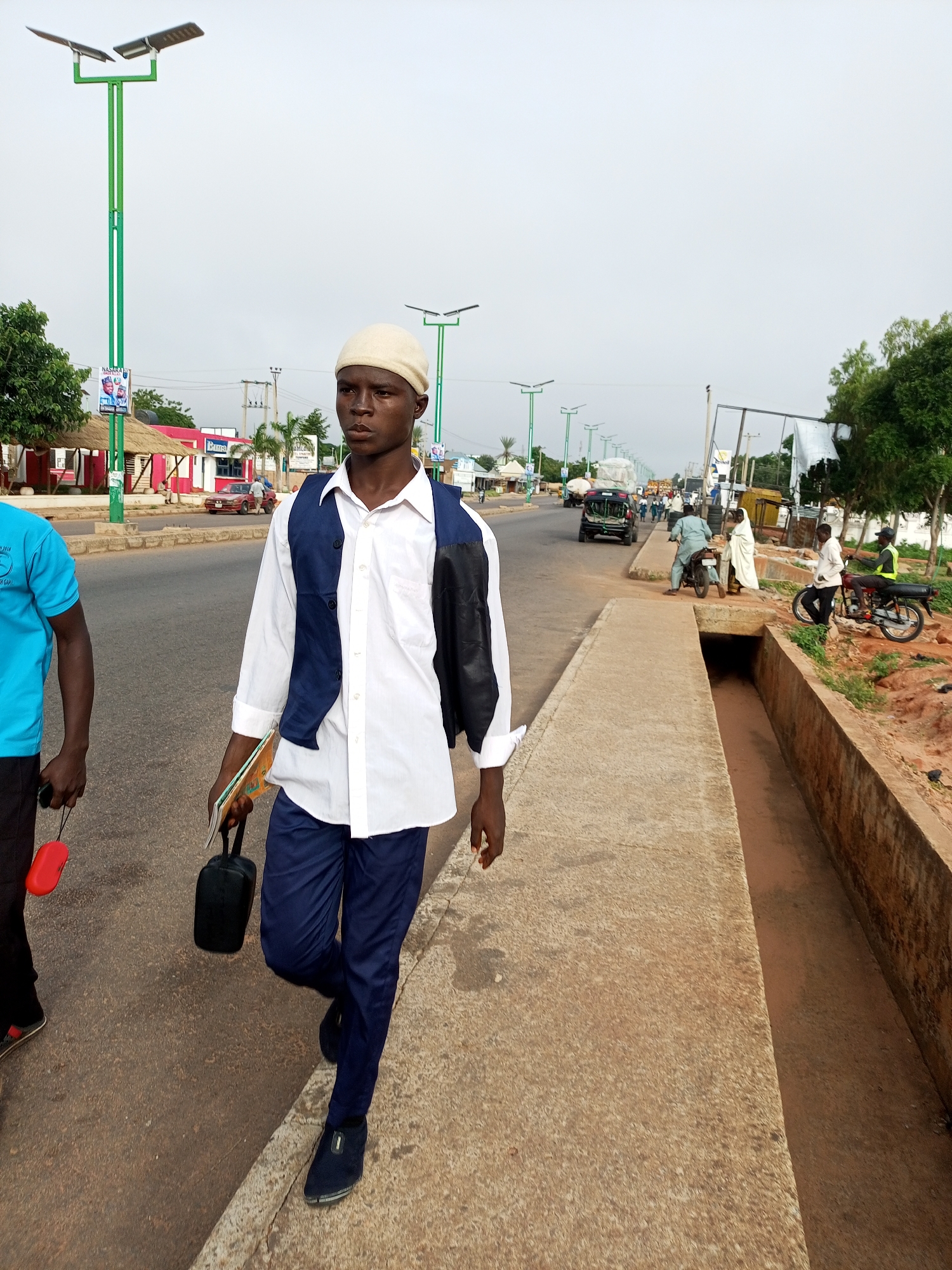 A young male wearing a white long-sleeved shirt, a dark blue vest, navy blue pants, and a light-colored cap walks along a paved sidewalk in Garko, Nigeria. He carries a black bag in his left hand and a book in his right hand, with a dark jacket draped over his right shoulder. To his left, another male wearing a blue shirt and black pants walks beside him. They are on a sidewalk next to a road with multiple lanes of traffic. In the background, there are solar-powered streetlights, buildings with thatched roofs, shops, and various vehicles, including cars, trucks, and motorcycles. Several other individuals are visible in the distance. The sky is overcast.