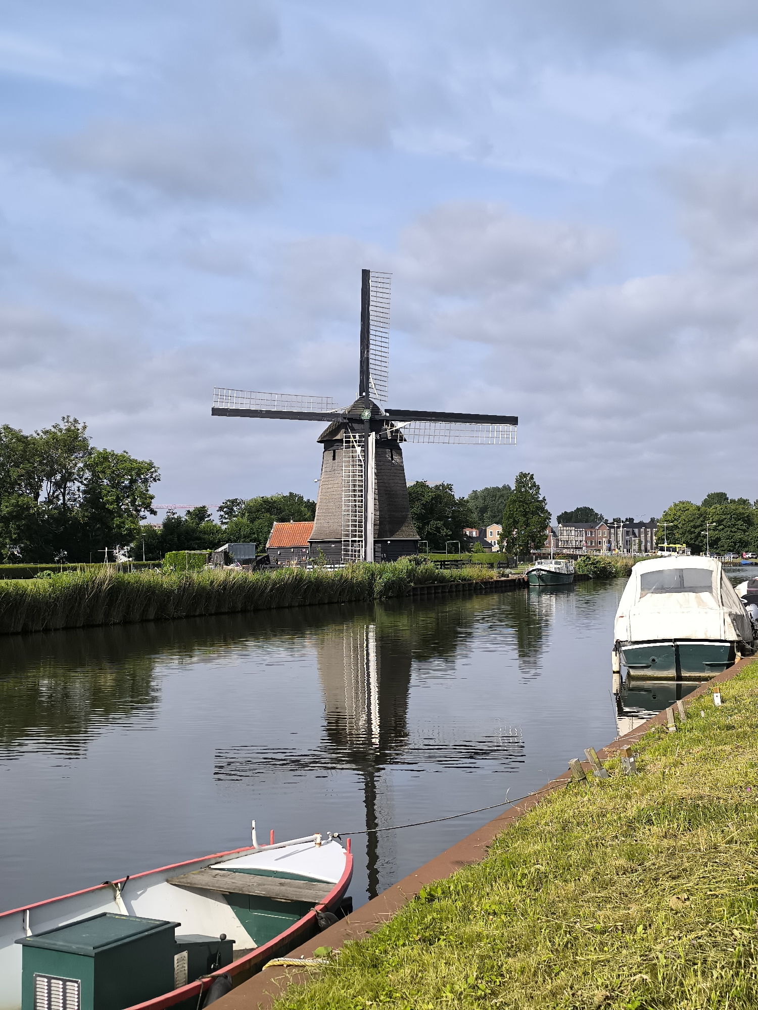A traditional Dutch windmill with its sails extended stands tall on the bank of a canal. The windmill, constructed of dark wood with a thatched roof, is a prominent feature of the landscape. A white and green motorboat is docked on the right side of the canal, covered by a white tarp. In the foreground, a smaller white and green boat with red trim is tied to the canal bank. The canal reflects the windmill, the sky, and the surrounding trees. The scene is set in Alkmaar, Netherlands, a city known for its historic windmills and canals. The sky is partly cloudy, with a mix of blue sky and scattered clouds, suggesting a pleasant day. Trees and buildings line the far bank of the canal, adding depth to the scene. The overall impression is one of peaceful, rural charm.