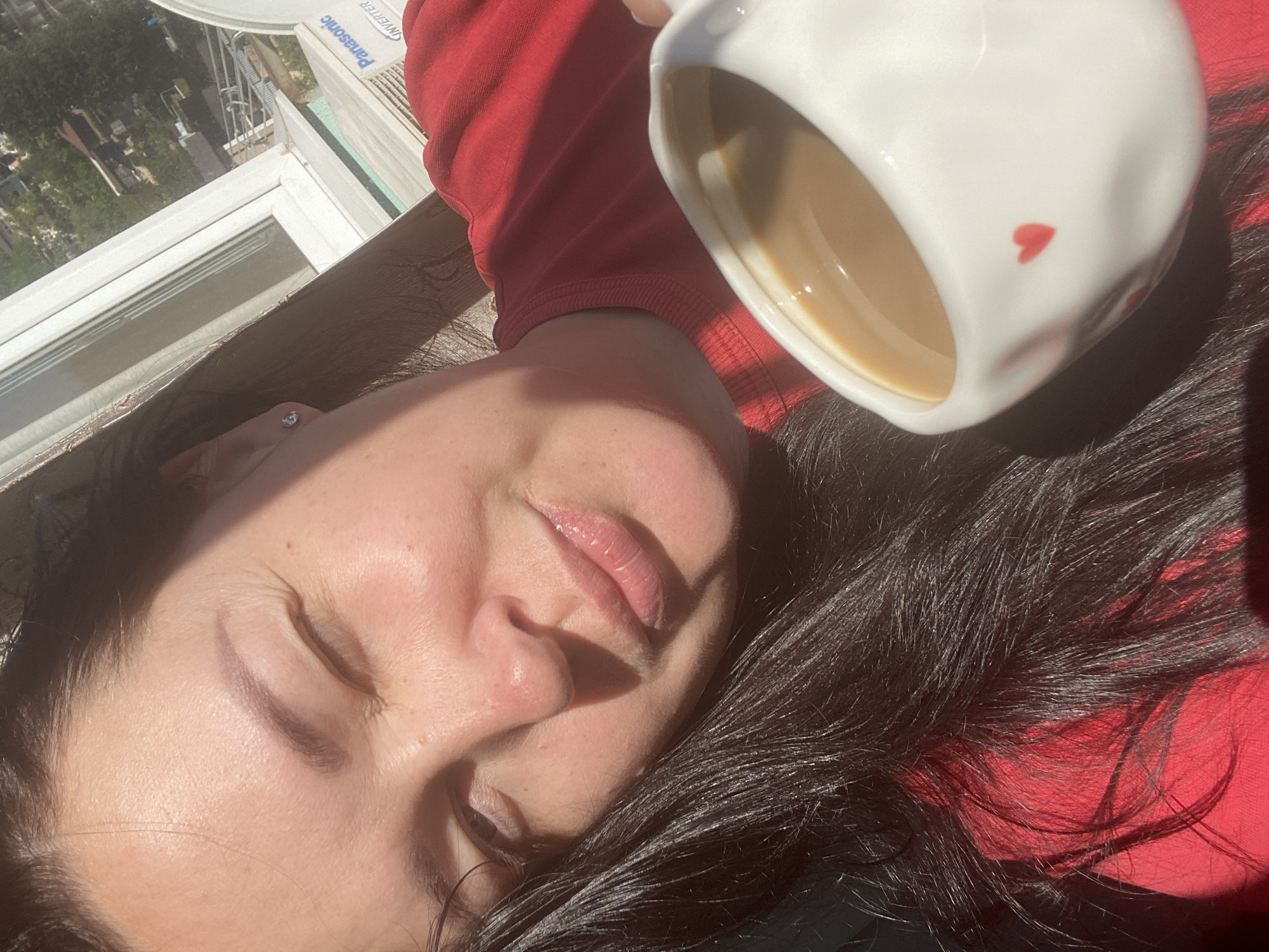 A woman with dark hair is lying down with her eyes closed. She is wearing a red shirt. A white mug with a red heart on it, filled with a light-colored liquid, is held above her head. In the background, a window and an outdoor air conditioning unit with "Panasonic Inverter" branding can be seen. The location is likely Sochi, Russia.