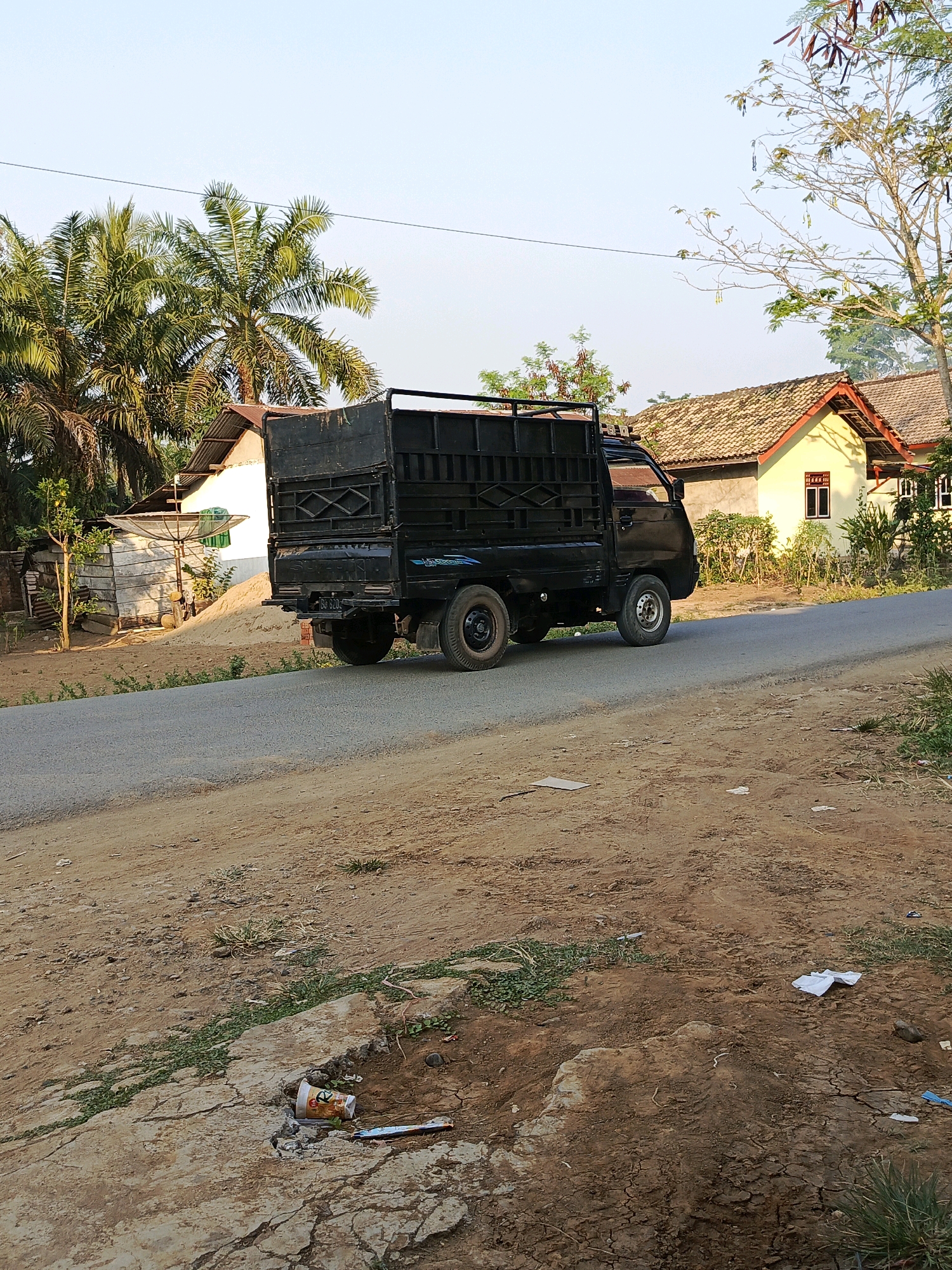 A black pickup truck with a cage bed is parked on the side of a paved road. The truck faces right, and its tires are visible. In the background, there are two houses with yellow walls and tiled roofs, surrounded by trees and vegetation. The sky is clear and pale blue. The ground in the foreground is dirt and cracked earth with some green grass growing through. There is some scattered litter on the ground. The scene is outdoors in what appears to be a rural or semi-rural area. The location is the city of Megang Sakti I, Indonesia.