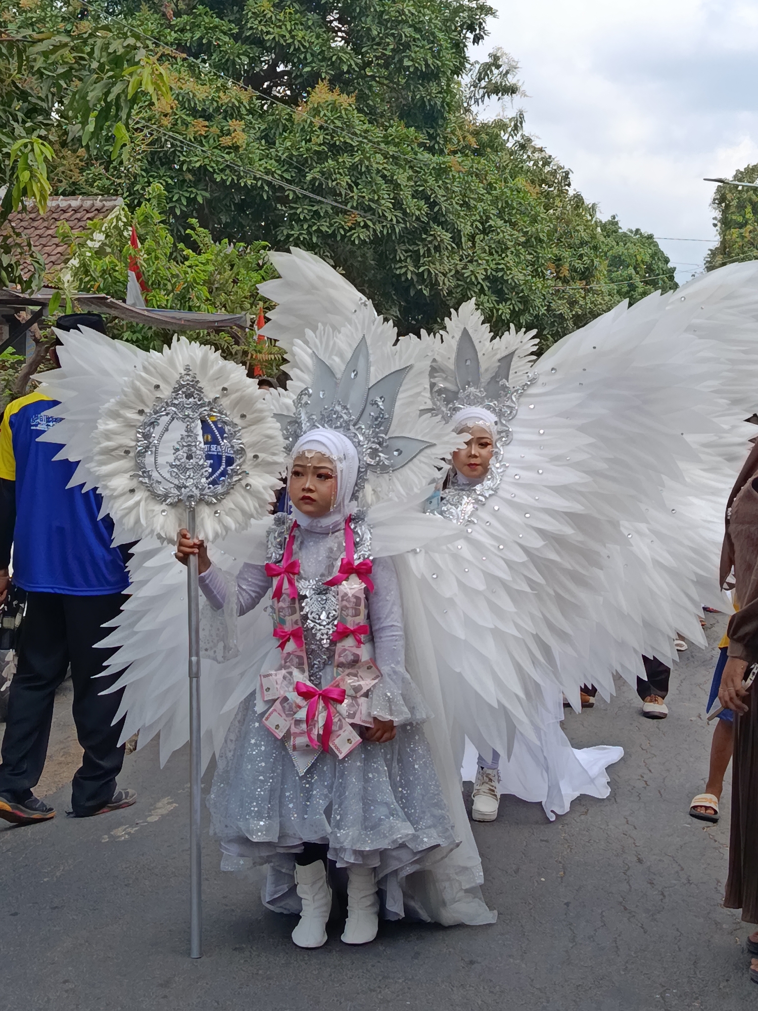 In Ketowan, Indonesia, two young girls dressed in elaborate white winged costumes participate in a parade. The girl in the foreground holds a large, ornate staff topped with a silver emblem. Her costume includes a shimmering silver dress and a white hijab, adorned with a necklace made of what appear to be banknotes tied with pink ribbons. She wears white boots. Beside her, a second girl, also in a white hijab and wings, is partially visible. The background features lush green trees and a cloudy sky. Other parade participants and spectators are visible in the periphery, with one person in a blue and yellow uniform to the left and others in casual attire walking on the paved street.
