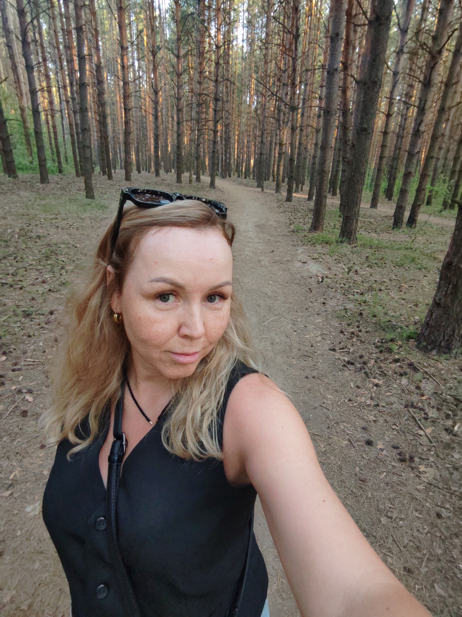A woman takes a selfie while walking on a dirt path through a dense pine forest. The forest has closely spaced, tall, slender trees lining both sides of the path. The woman has blonde hair, sunglasses perched on her head, and wears a dark sleeveless top. She's looking at the camera with a slight smile. The ground is covered in pine needles and leaf litter. The location is the city of Chaikovsky, Russia.