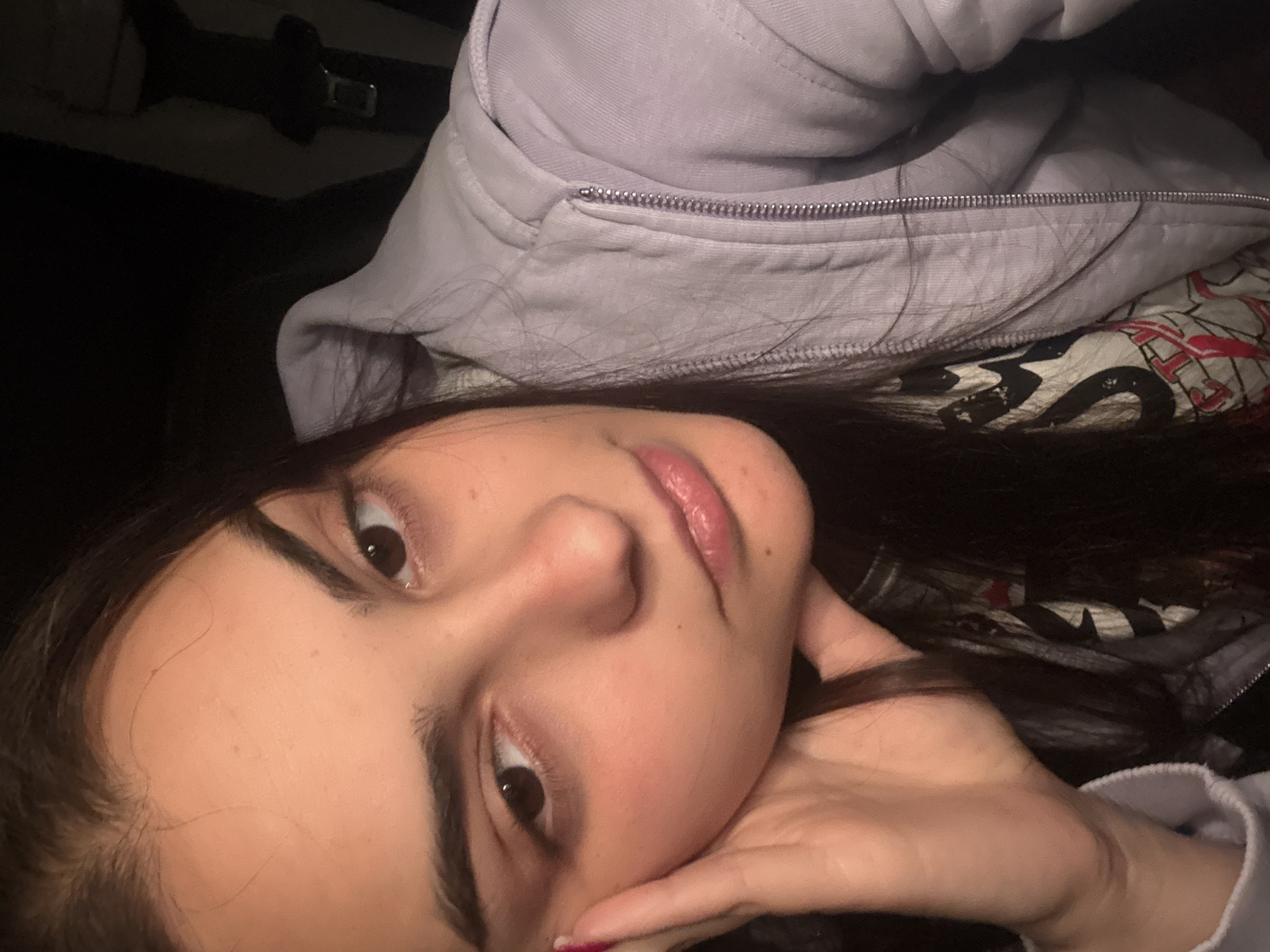 A young woman is depicted in a close-up, tilted selfie. She has fair skin, dark brown hair, and brown eyes. Her face is partially supported by her left hand. She is wearing a light purple hoodie with a visible silver zipper. Behind her is a patterned fabric with white, red, and black designs. The overall lighting is dim, and the background is dark. There is no indication of the location being Elenovo, Bulgaria.
