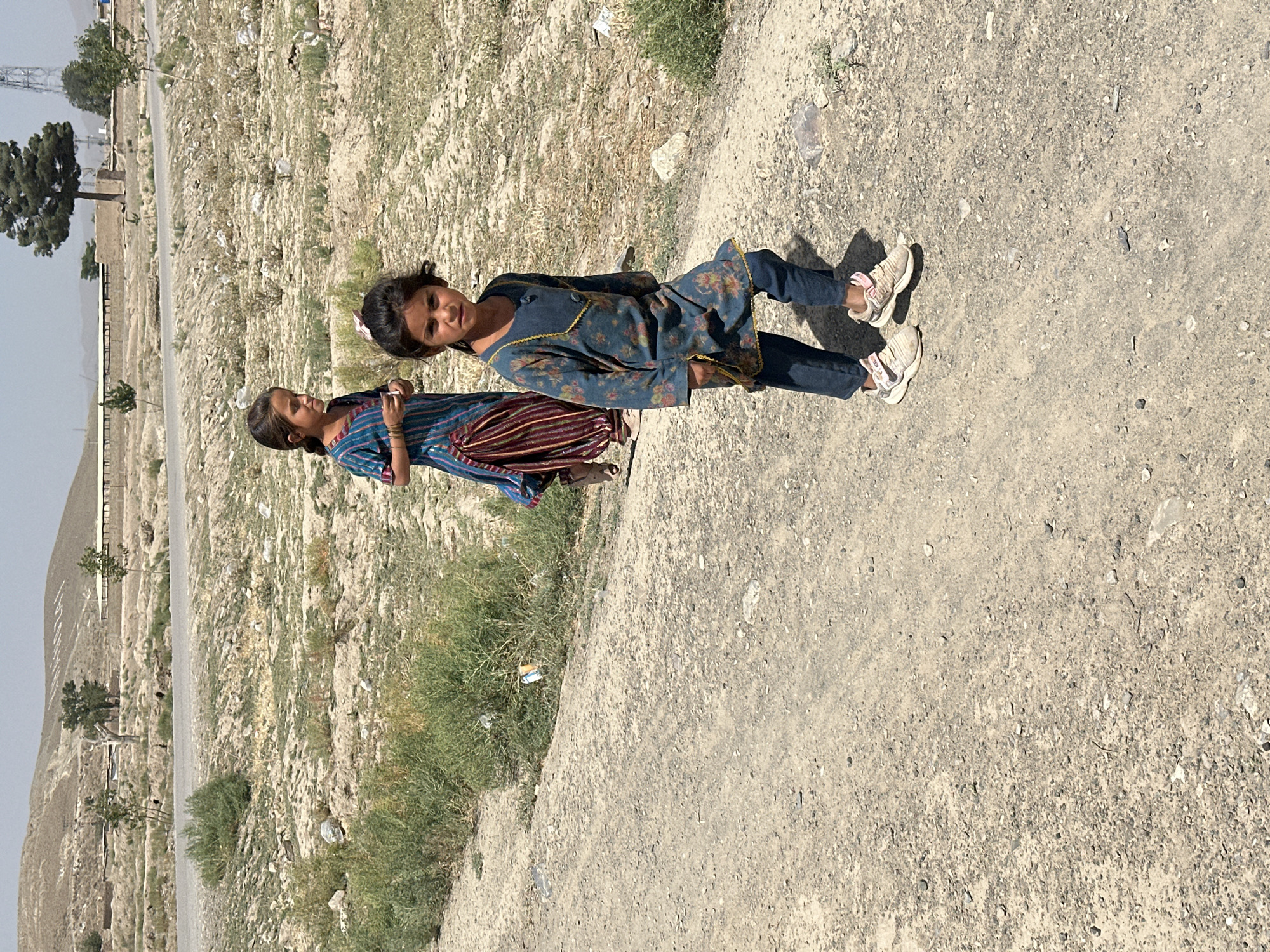 In Kanozai, Pakistan, two young girls walk on a dirt road. The girl in the foreground wears a blue floral patterned dress over dark blue pants and white sneakers. The girl behind her is dressed in a blue and striped traditional outfit. The environment is arid, with dry vegetation and sparse trees visible in the background. A road is present in the mid-ground. The sky is clear and bright, suggesting a sunny day.