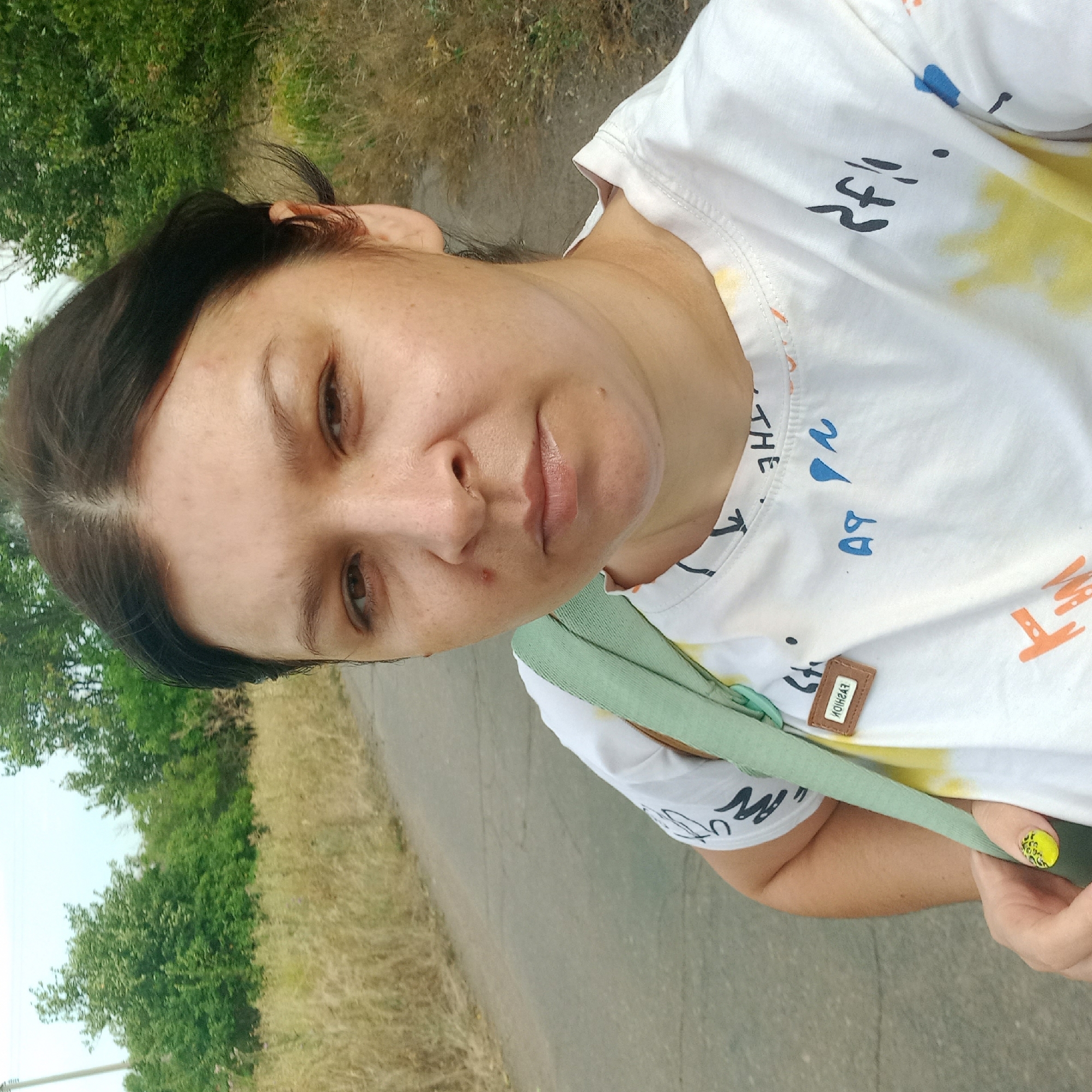 A woman is taking a selfie on a paved road lined with dry grass and trees. She has dark hair, brown eyes, and is wearing a white t-shirt with colorful abstract designs and a light green strap across her shoulder. Her left hand, adorned with a yellow and black patterned nail, is visible. A small brown tag with the word "FASHION" is attached to her shirt. The background suggests a rural or suburban environment with a power pole visible in the distance on the left. The lighting indicates it is daytime.