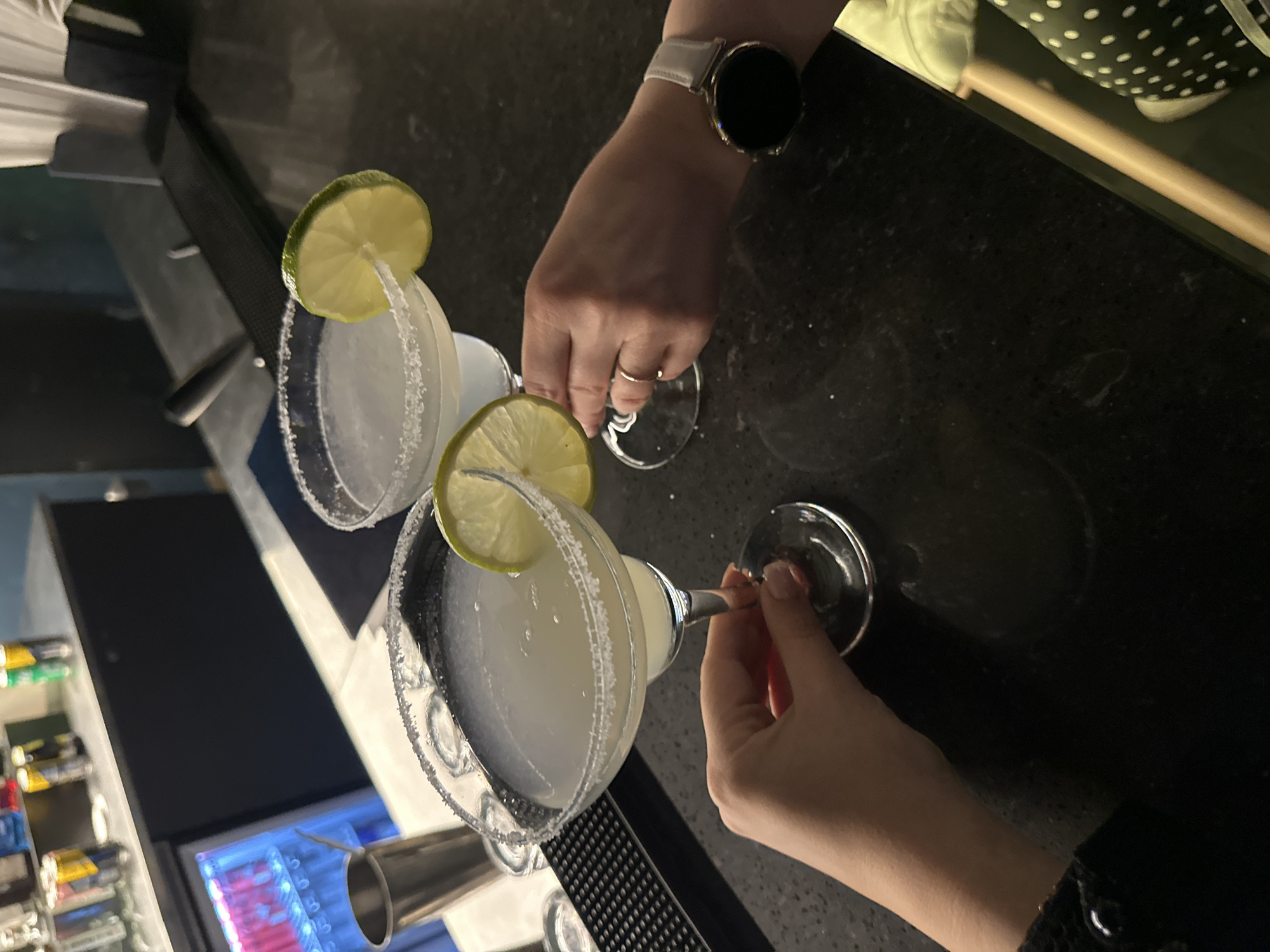 The image, taken from an overhead perspective, depicts a bar scene featuring two individuals holding margarita cocktails. Two glasses, each rimmed with salt and garnished with a fresh lime wedge, are prominent on a dark, speckled bar counter. The drinks themselves appear to be a light, opaque liquid, typical of margaritas.
One hand, wearing a white smartwatch with a circular face and a simple ring on the ring finger, holds the stem of the upper margarita glass. Another hand, with light pink nail polish, is seen holding the stem of the lower glass. The composition suggests a social interaction, possibly a toast or a moment before enjoying the drinks.
The setting appears to be an indoor bar or restaurant, given the presence of a dark bar counter and bar equipment, including a metallic jigger and a black bar mat visible in the lower-left corner. In the background, a commercial refrigerator with illuminated shelves displays an assortment of cans or bottles, though their specific labels are indistinct. The dim lighting implies an evening or nighttime ambiance. No specific text is legible in the image. The scene conveys a relaxed, social atmosphere.