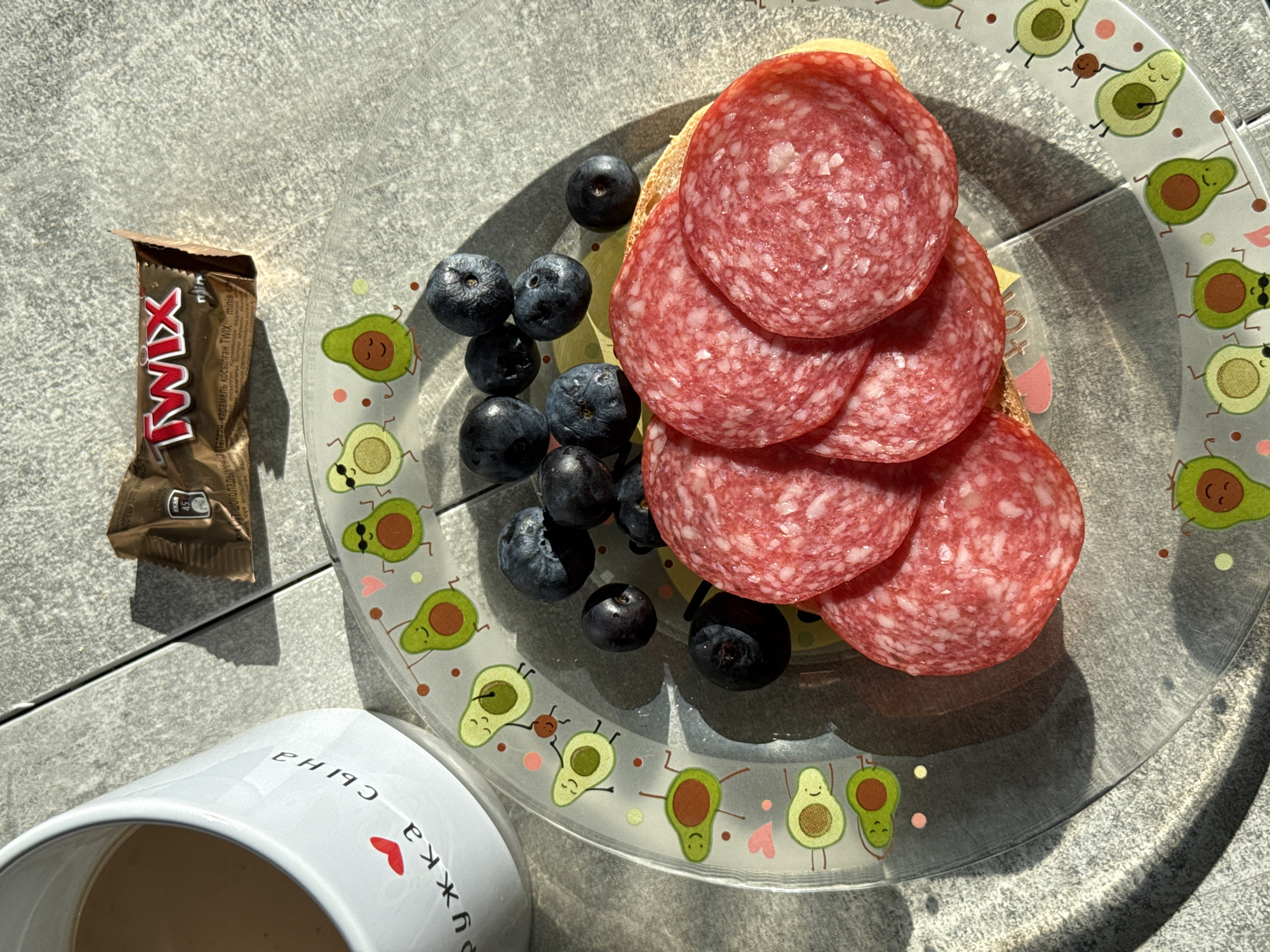 A close-up overhead shot shows a breakfast spread on a textured gray surface, likely a table outdoors. In the upper left corner, a white ceramic mug with a red heart and black Cyrillic text is partially visible. To the right of the mug, a small, unwrapped Twix chocolate bar lies on its side. The main focus of the image is a clear, round plate decorated with cartoon avocados wearing sunglasses and dancing. On the plate are several slices of salami arranged on top of a piece of bread, accompanied by a cluster of fresh blueberries. The lighting suggests a sunny day, casting distinct shadows across the scene. The scene is set in Lyubertsy, Russia.