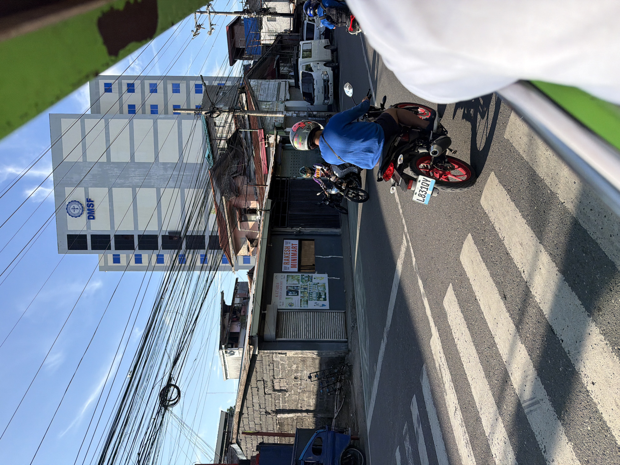 In Davao City, Philippines, a wide, sun-drenched asphalt road features a pedestrian crossing marked by white stripes. Several motorcycles navigate the street, with one in the foreground bearing the license plate L831QY. Behind the motorcycles, a multi-story building with a gray facade and blue windows displays the logo "DMSF." Adjacent to the building is a row of smaller establishments, including "RAKESH MINIMART," characterized by a metallic roll-up door and signage. The sky above is a clear blue, dotted with wispy white clouds. Overhead, a dense network of electrical wires crisscrosses the sky, a common sight in urban Philippine landscapes. The image appears to be taken from within a moving vehicle, evidenced by the blurred green border in the foreground and the reflection on the vehicle's side.