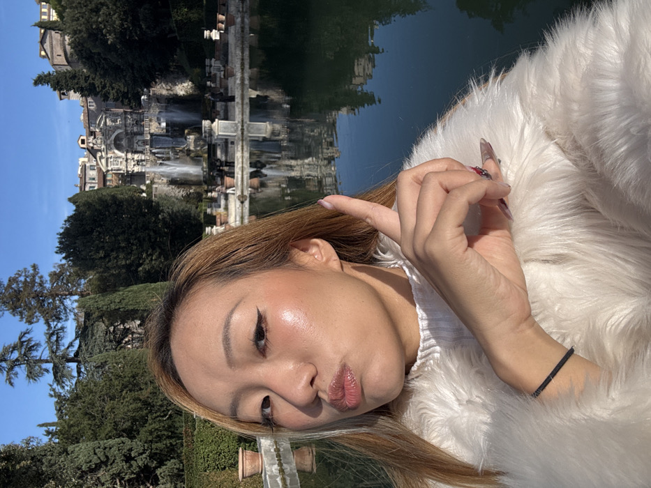 The image is a selfie depicting an East Asian female subject in the foreground, positioned in the gardens of Villa d'Este in Tivoli, Italy. The subject has long, light brown hair and is wearing a white ribbed knit top under a white faux fur jacket. Her face is turned towards the camera with lips pursed, and her right index finger is extended upwards. Visible makeup includes winged eyeliner and glossy lips; her fingernails are long and styled, with a patterned design on the ring finger. A black elastic band is present on her left wrist.
In the background, a large, dark reflecting pool occupies the lower portion of the frame, showing reflections of the distant architecture and trees. Beyond the pool, the multi-tiered Fontana del Nettuno (Fountain of Neptune) is prominently featured, actively discharging water. The fountain is surrounded by dense green foliage, including tall cypress trees and manicured hedges. Buildings are visible on the hillside above and behind the fountain structure. Several indistinct human figures are discernible on the pathway in front of the fountain. The scene is brightly illuminated by natural sunlight under a clear blue sky.
