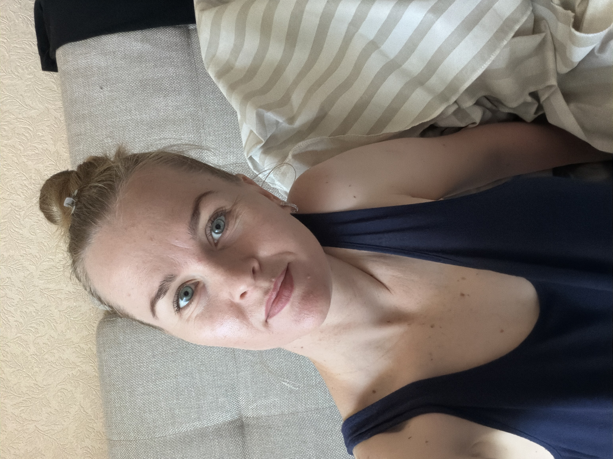 A woman with blonde hair pulled into a bun and blue eyes is looking directly at the camera. She is wearing a dark blue tank top. The background appears to be a bed with a textured wallpaper. There is a striped duvet or comforter to her right. The lighting suggests it is daytime. There is no indication of the location in Khabarovsk, Russia.