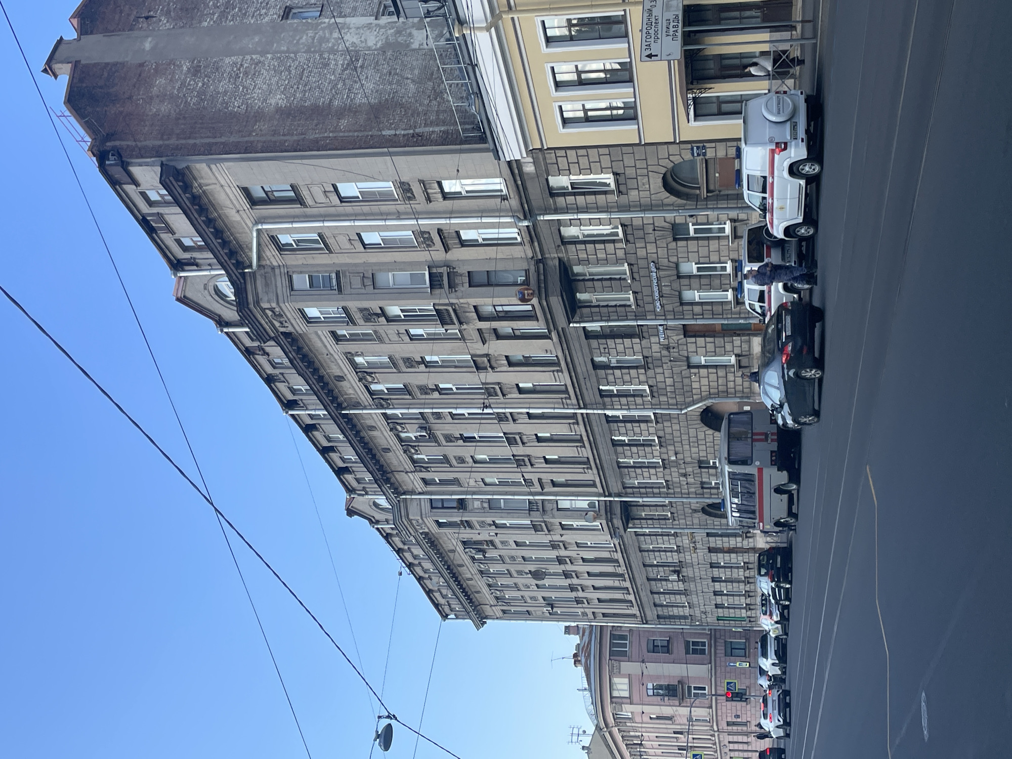 The image shows a street scene in Saint Petersburg, Russia, on a clear day. In the foreground, a dark asphalt road with lane markings is visible. Several cars, including a dark sedan, a white SUV with red accents, and another white SUV, are parked on the side of the road. Two individuals, appearing to be law enforcement officers in uniform, stand near the white SUVs. A grey bus with red stripes is also parked along the street.
Dominating the background is a large, multi-story building with a grey stone facade, characterized by numerous windows and ornate architectural details. To the right of this building, a smaller yellow building with a white trim and windows is visible. A street sign on the yellow building indicates directions to "ЗАГОРОДНЫЙ ПРОСПЕКТ" (Zagorodny Prospekt) and "УЛИЦА ПРАВДЫ" (Ulitsa Pravdy). Further to the left, another building with a pink facade and rounded architectural elements can be seen. Overhead, power lines crisscross the blue sky. The scene appears to be a typical urban environment with no immediate signs of distress or unusual activity.